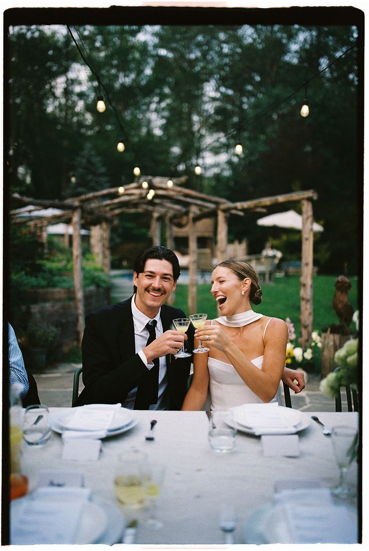 A couple shares a joyful laugh during an outdoor reception dinner under string lights and wooden posts.
