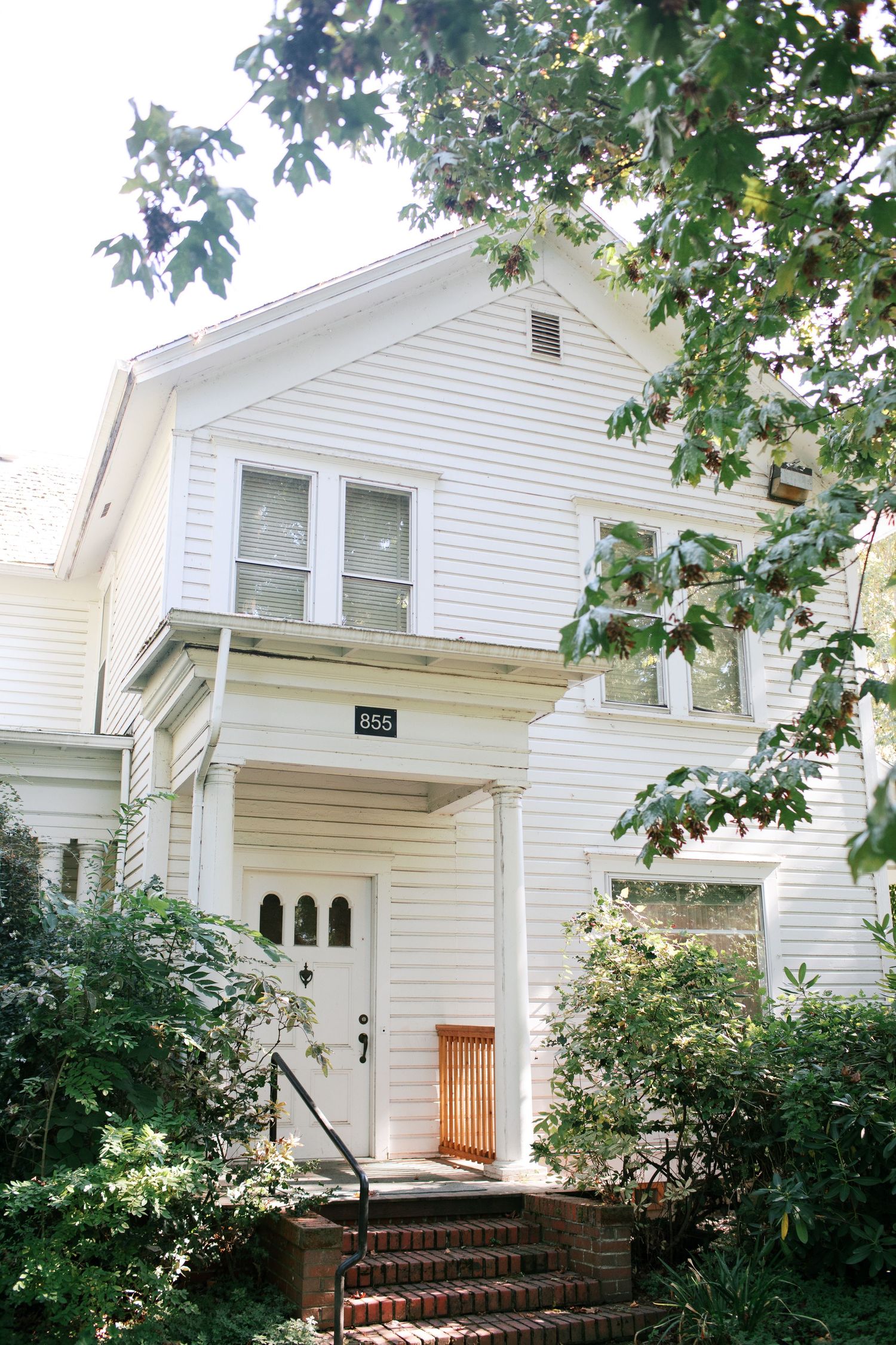 White wooden Victorian-style house exterior with front porch steps and leafy tree branches framing the view.