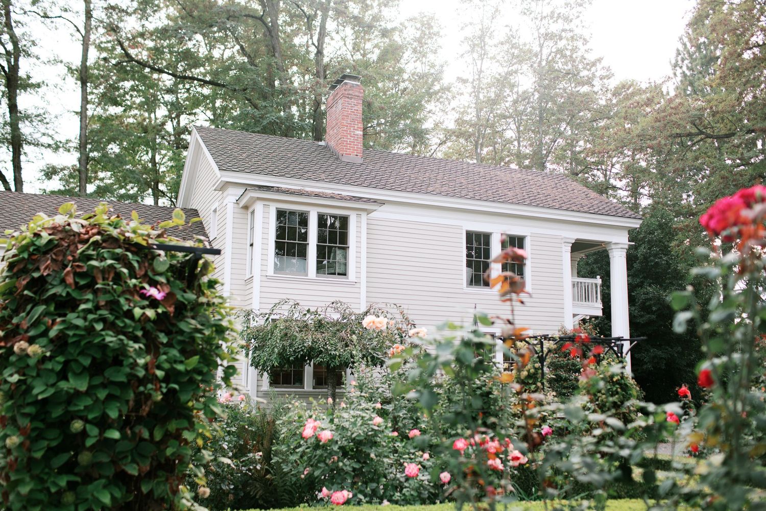 A charming white cottage house with a grey roof is nestled among lush green trees and flowering gardens.