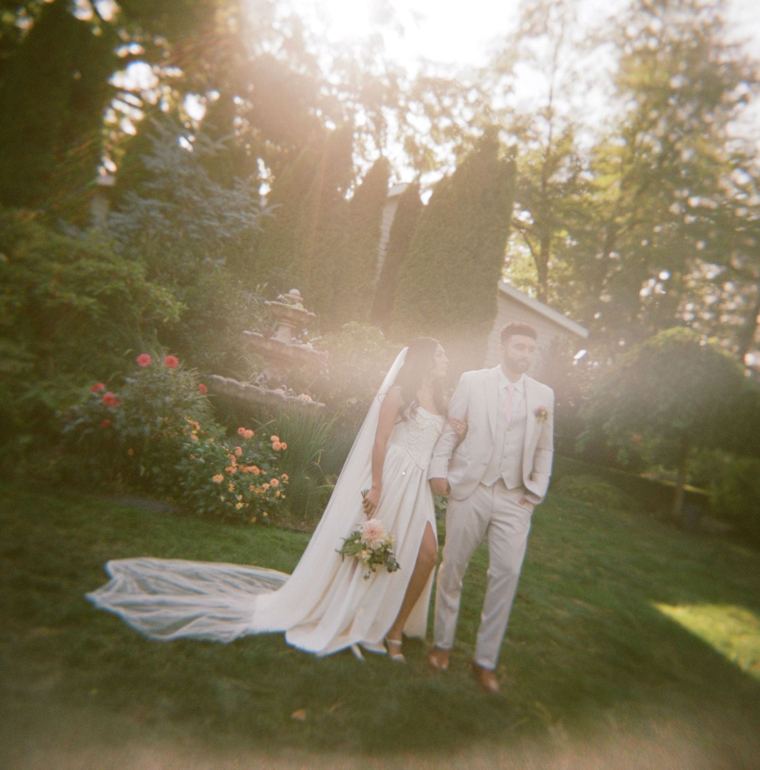 A romantic wedding couple poses in a sunlit garden setting during golden hour with flowing veil and white attire.