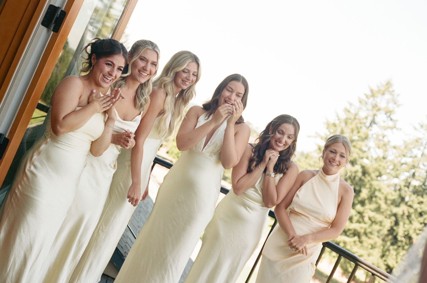 A group of bridesmaids in elegant cream-colored dresses share a joyful moment together on a balcony.