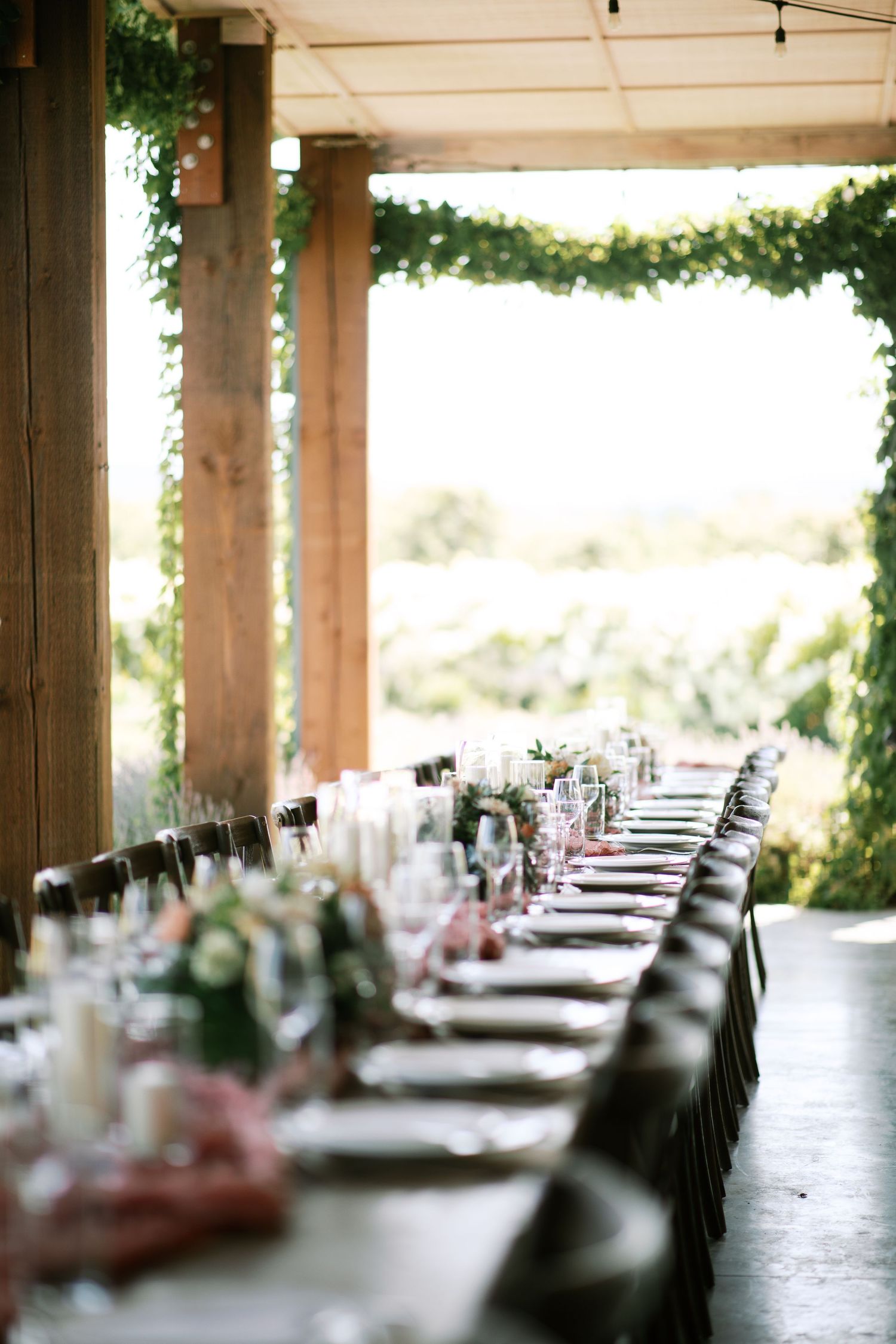 A long dining table set for an outdoor wedding reception under a covered patio with natural lighting and greenery.