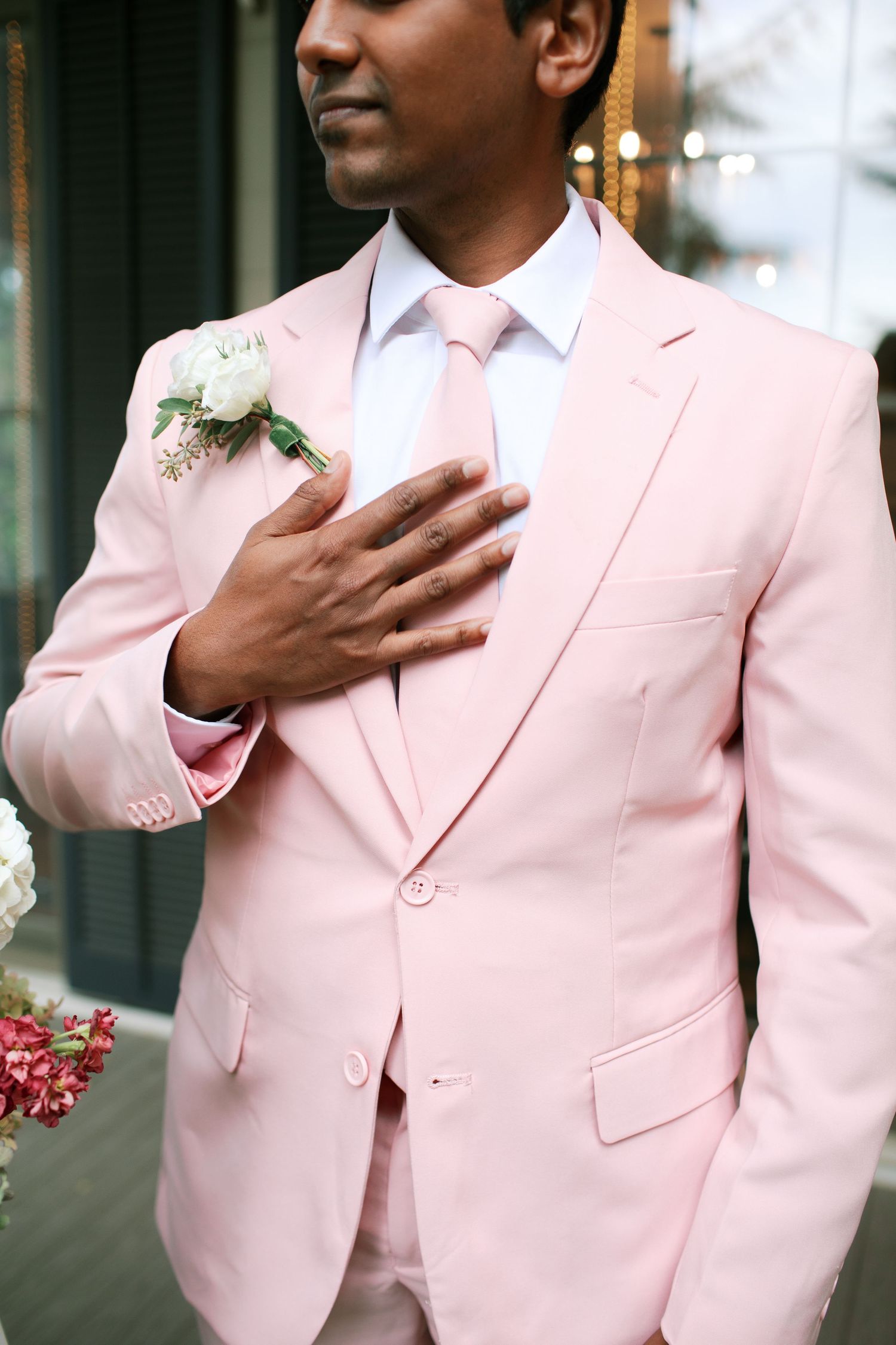 A person wearing a light pink suit jacket with a white boutonniere poses with their hand over their heart.
