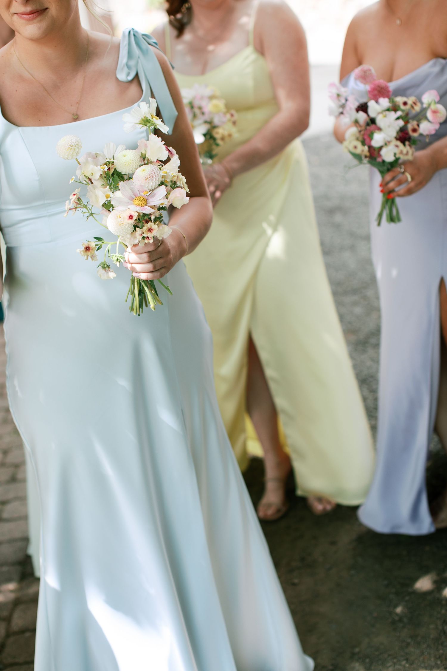Bridesmaids in pastel yellow and light blue dresses hold white floral bouquets at an outdoor wedding.