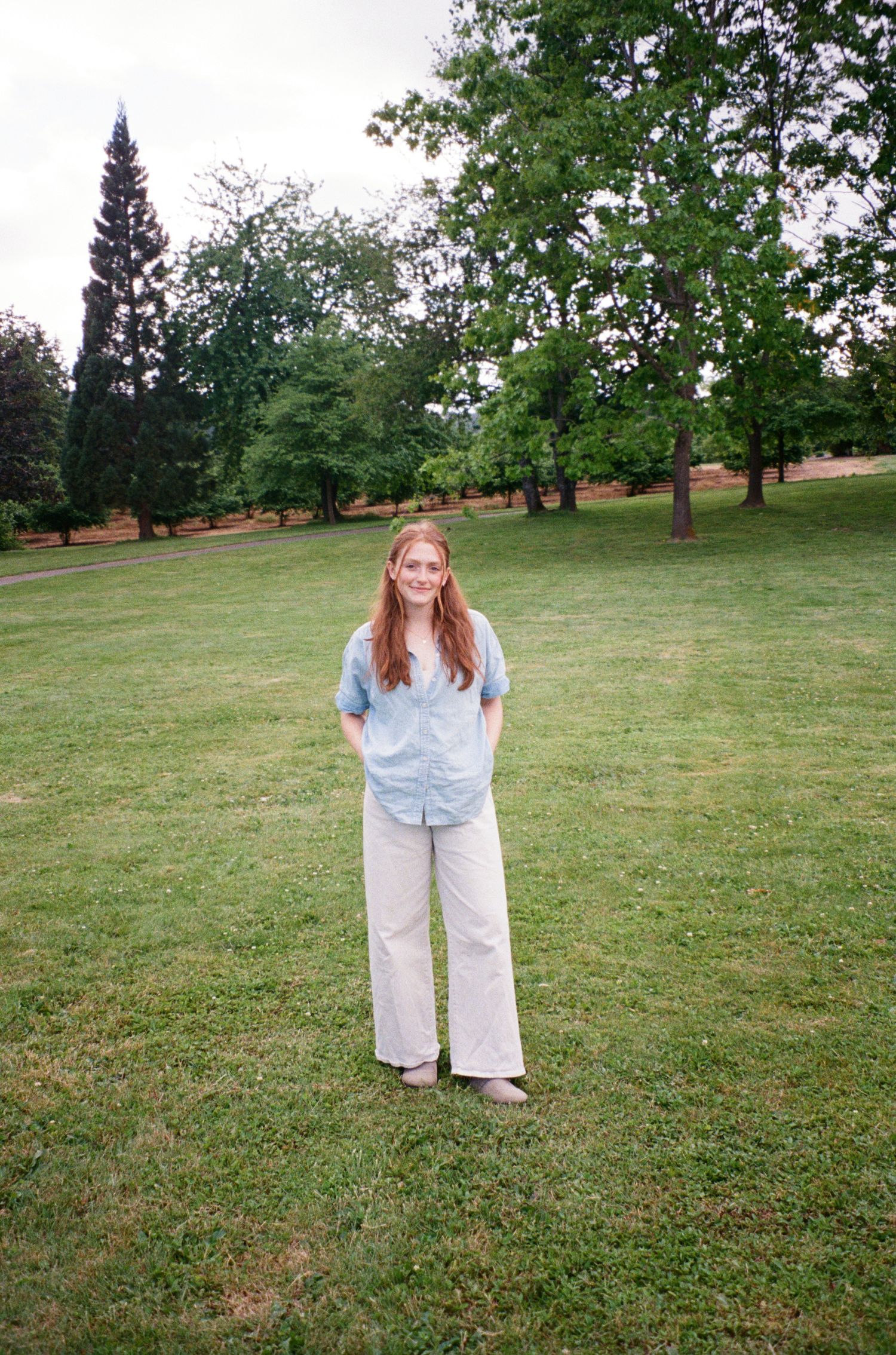A person wearing a light blue shirt and white pants stands in a grassy field surrounded by trees on a sunny day.