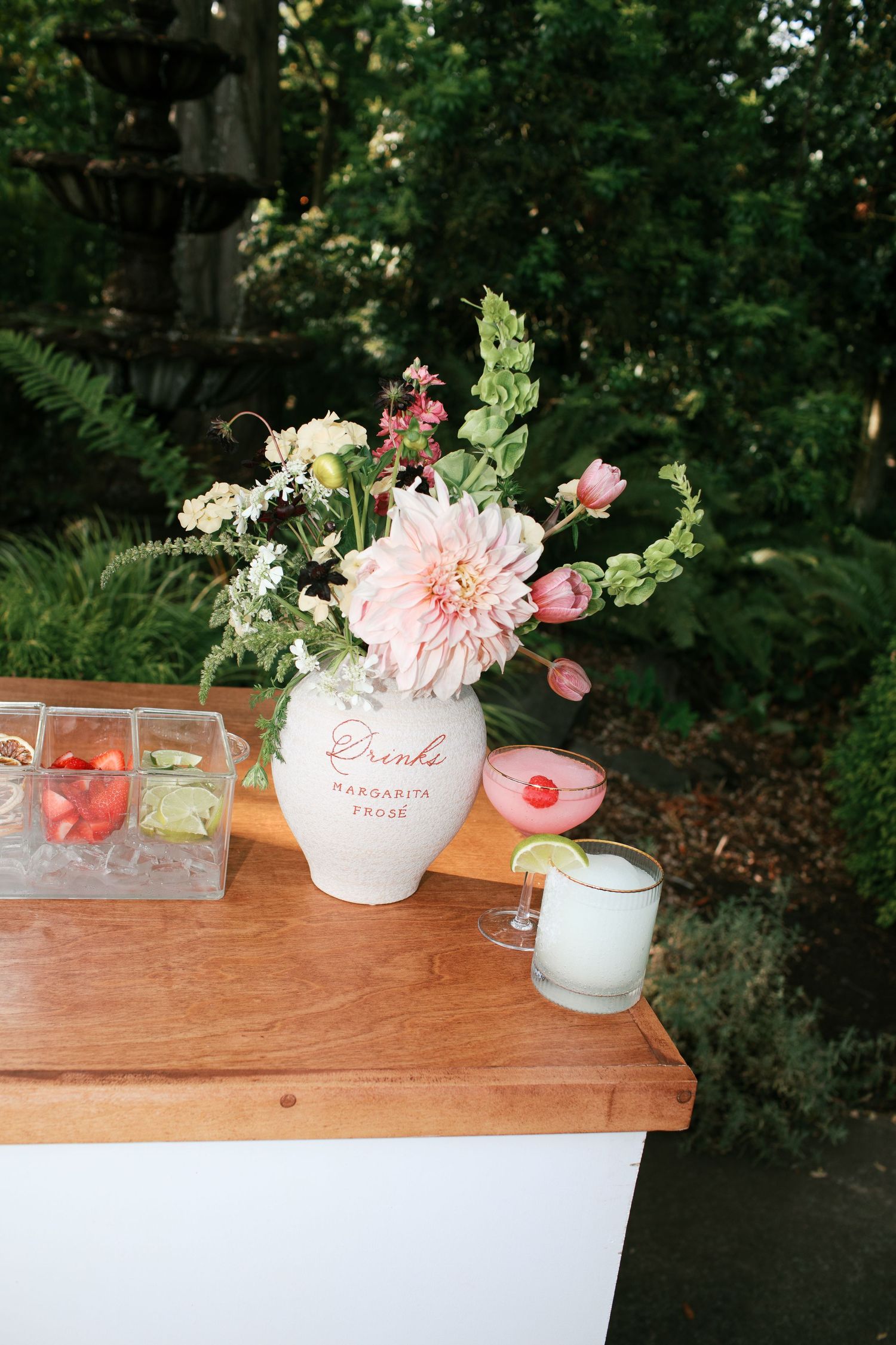 Pink and white floral arrangements in vintage vases sit atop a rustic wooden table against a lush garden backdrop.