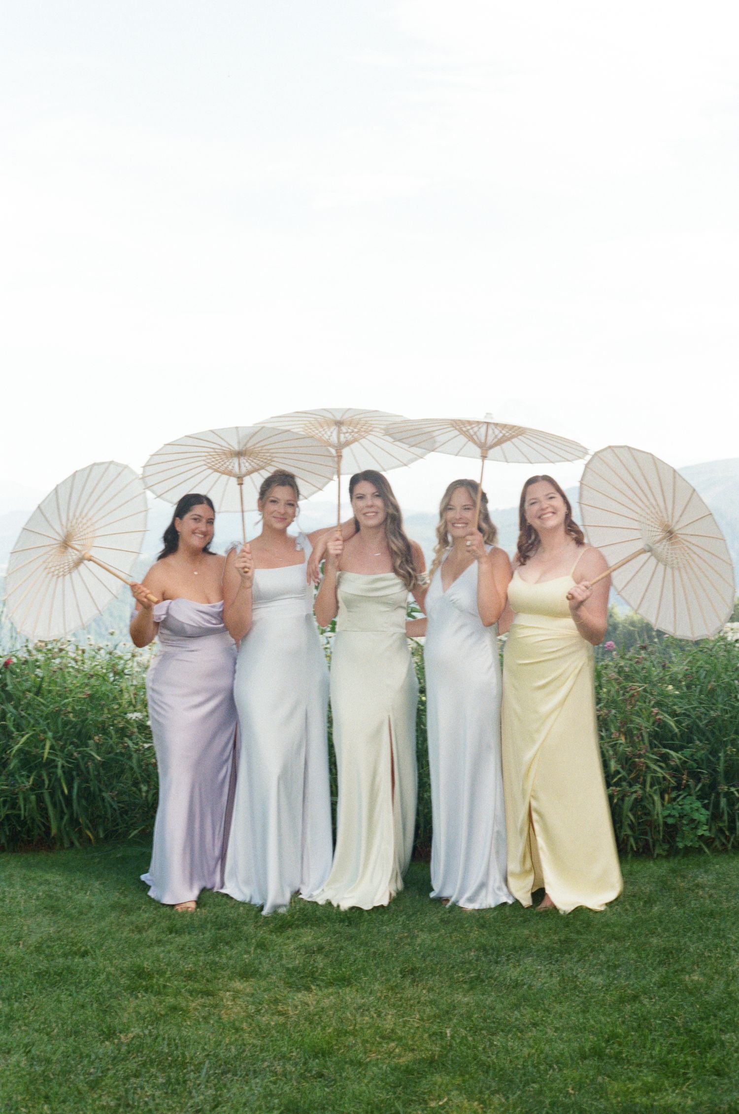 Bridesmaids in pastel dresses pose together holding white paper parasols on a grassy lawn.