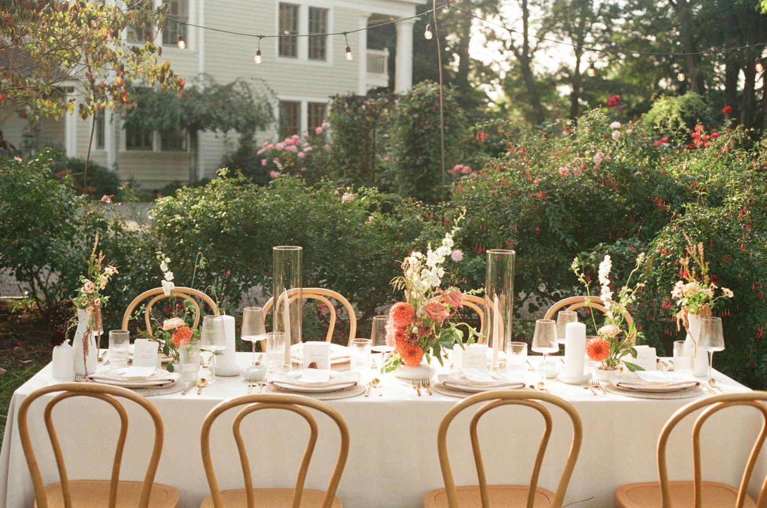 Elegant outdoor wedding reception tables with wooden chairs and floral centerpieces set against a garden backdrop.