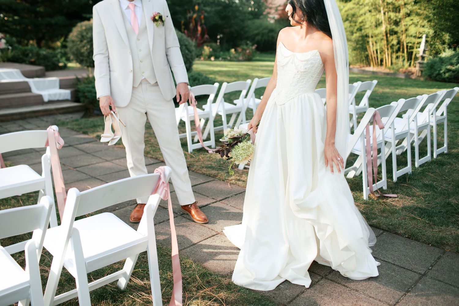 A couple in wedding attire walks along a stone path lined with white folding chairs decorated with pink ribbons.