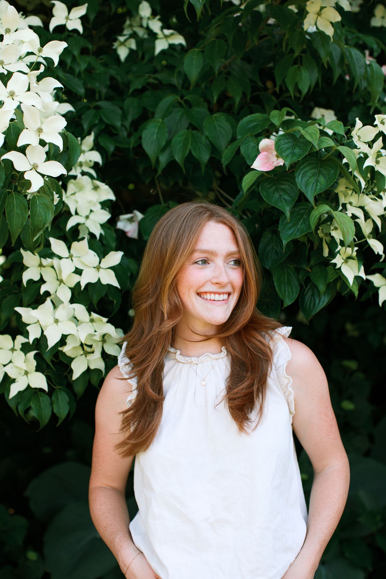Smiling woman with long auburn hair in white sleeveless top standing before lush green foliage with white blooms.