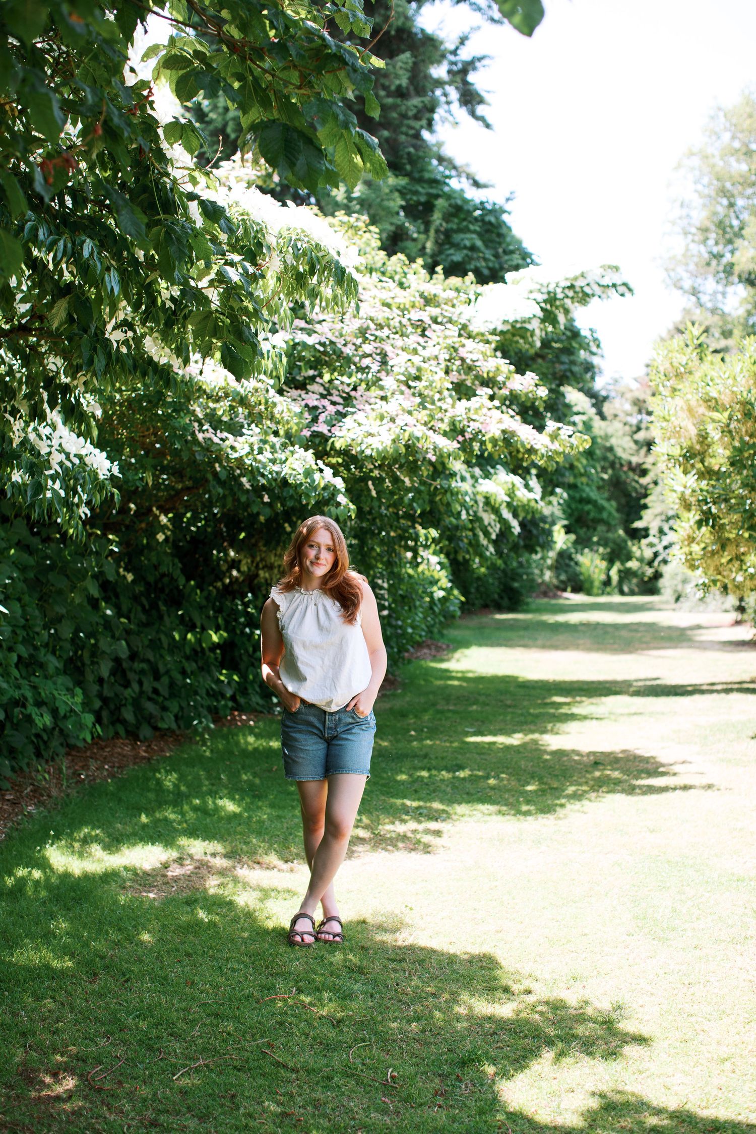 A young woman in white top and denim shorts walks along a lush green garden path lined with flowering trees.