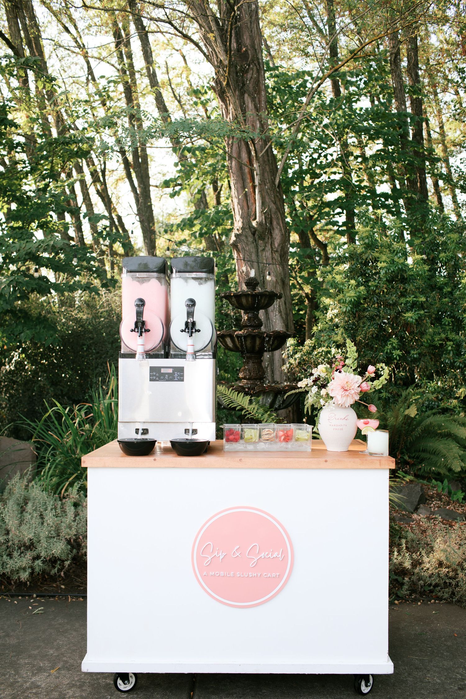 A white mobile beverage cart with pink logo features two drink dispensers and floral decor in an outdoor garden setting.