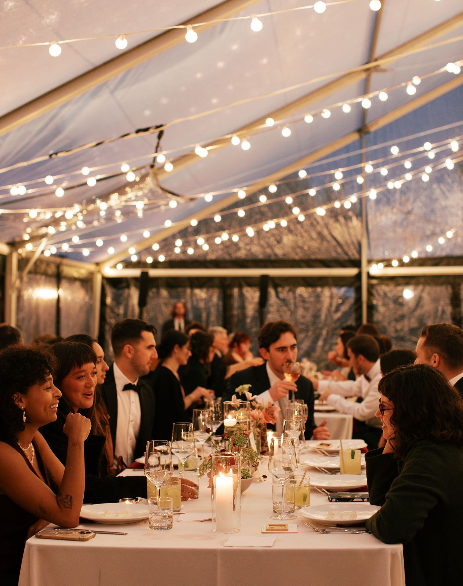Guests seated at elegantly decorated tables under string lights in a wedding reception tent at dusk.