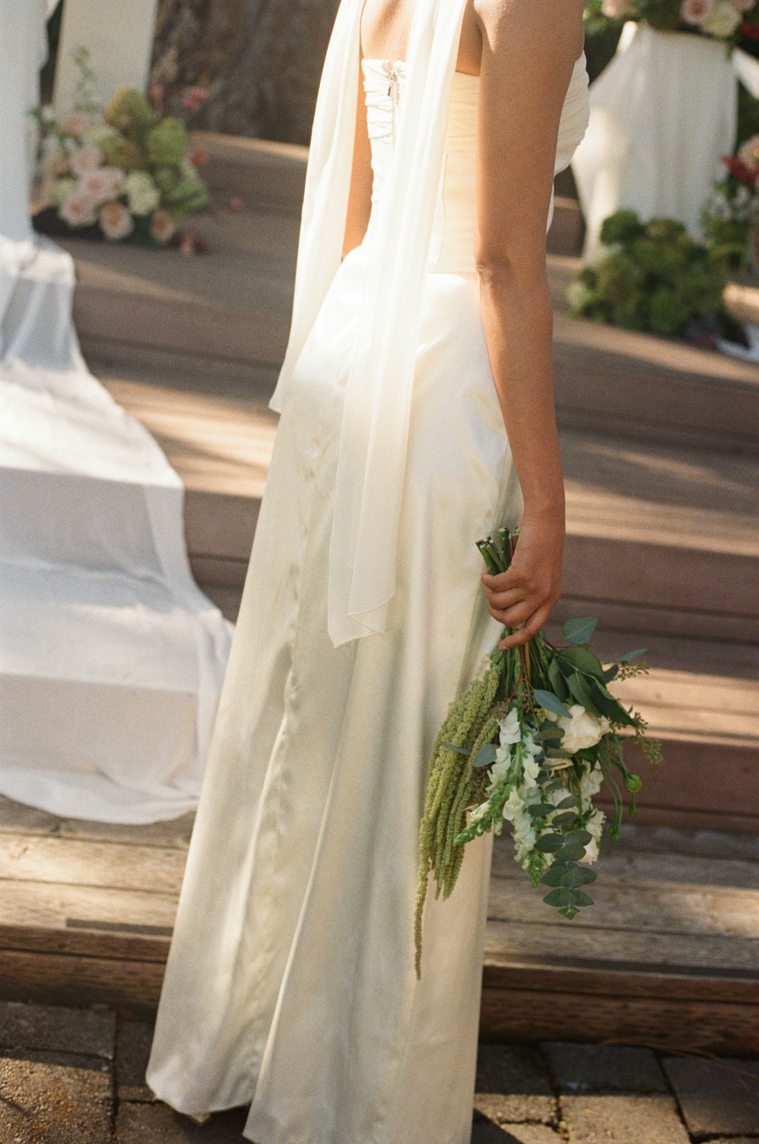 A flowing white wedding dress with a delicate bouquet of green stems and white flowers held at the side on stone steps.
