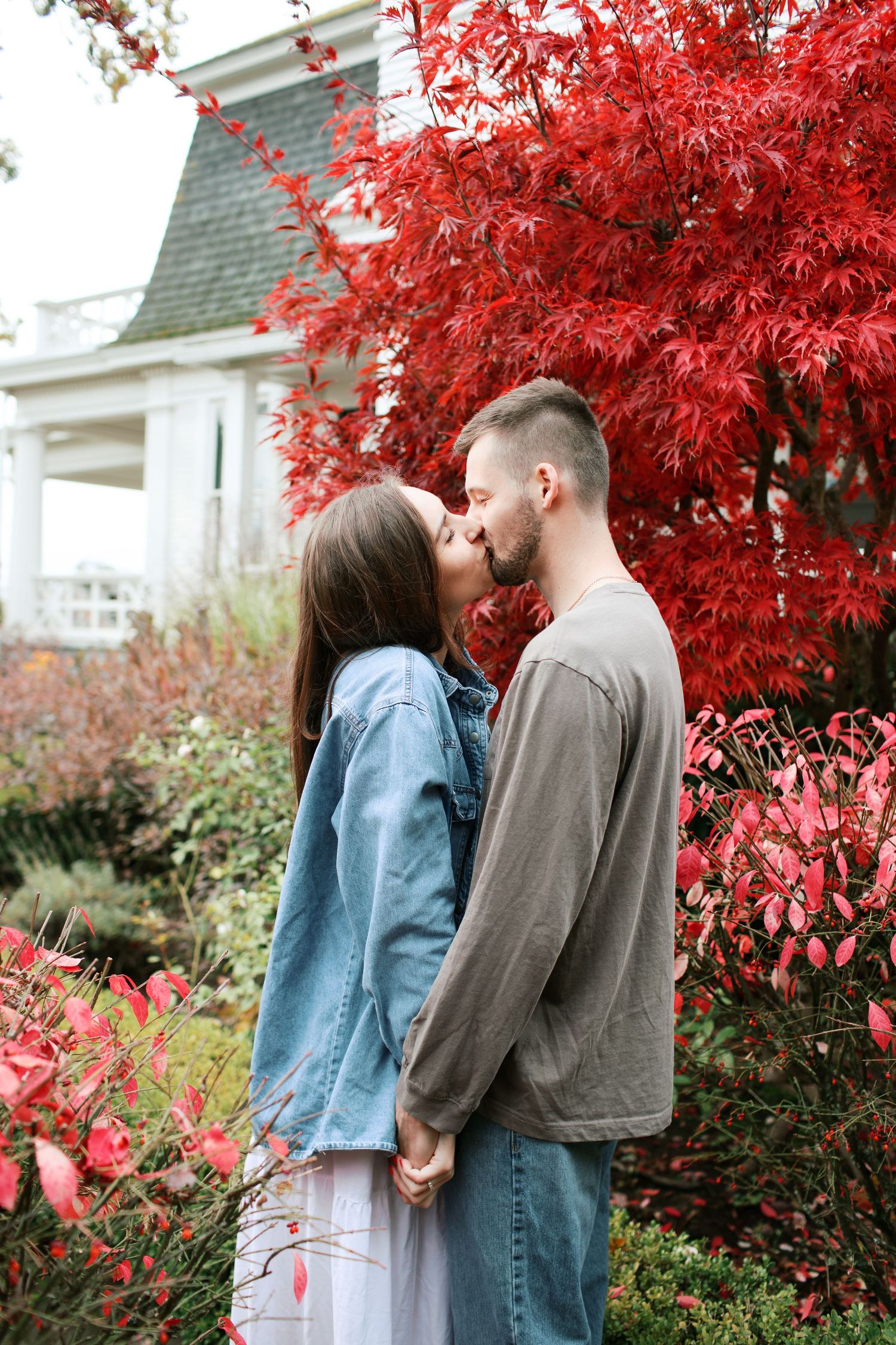 A couple embraces under vibrant red autumn maple leaves beside a white house exterior.