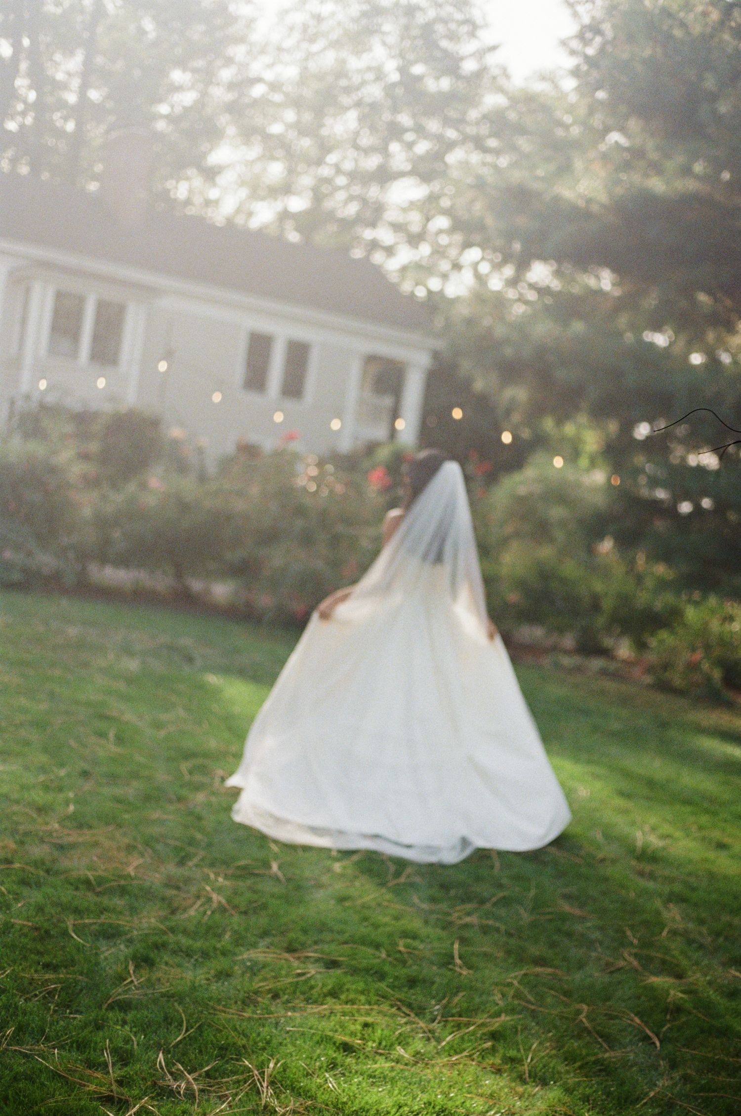 A person in a flowing white wedding dress and veil stands on a sunlit lawn with a house in the background.