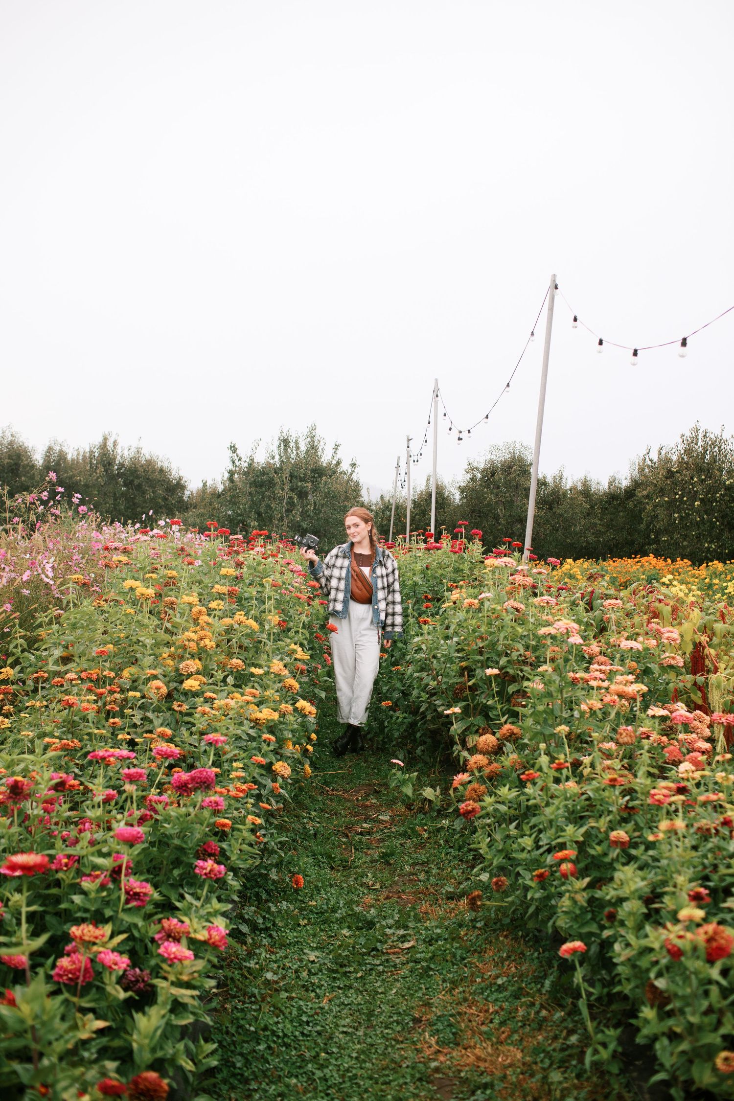 Adrianna Elizabeth photography walks through a vibrant garden path filled with colorful dahlias and wildflowers on a cloudy day.