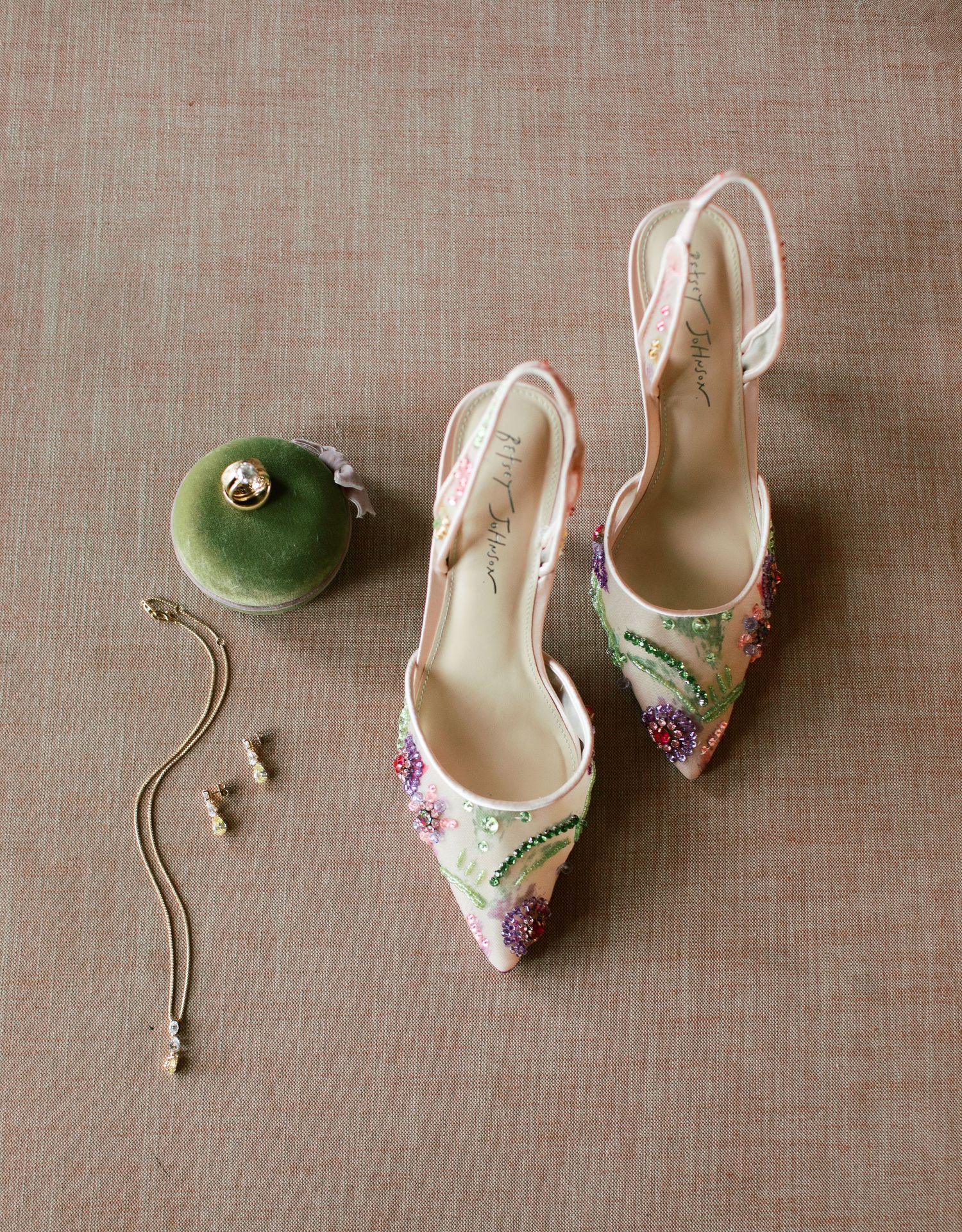 Elegant floral embroidered slingback heels displayed with a green ornament and delicate jewelry on linen background.