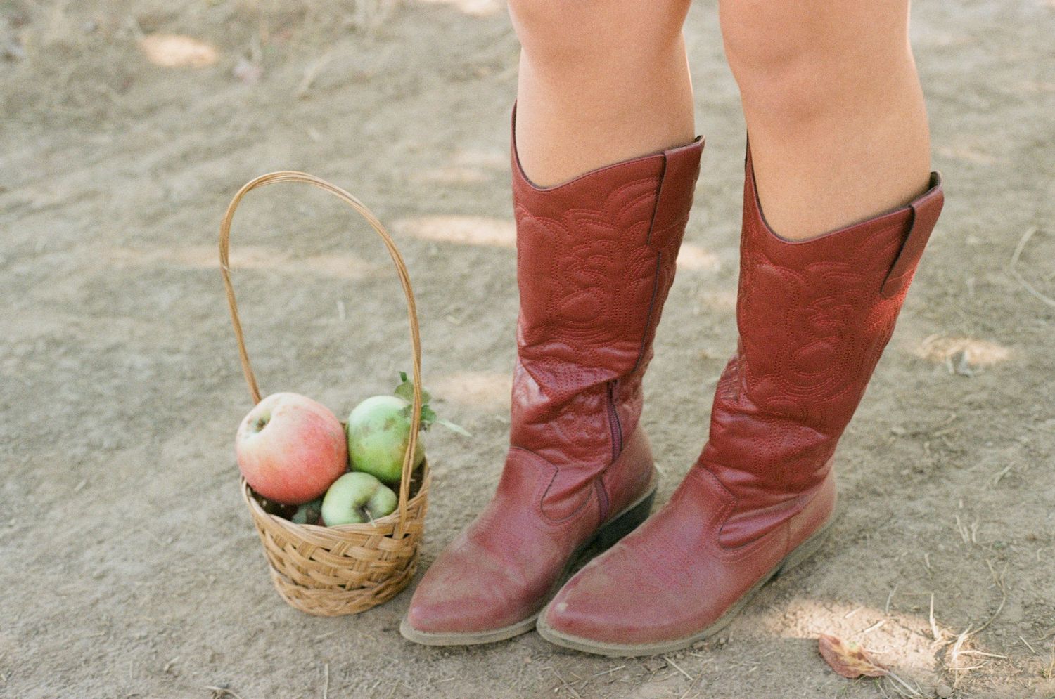 Red leather boots stand next to a small woven basket with apples on a dusty ground on 35mm film-Adrianna Elizabeth Photography.