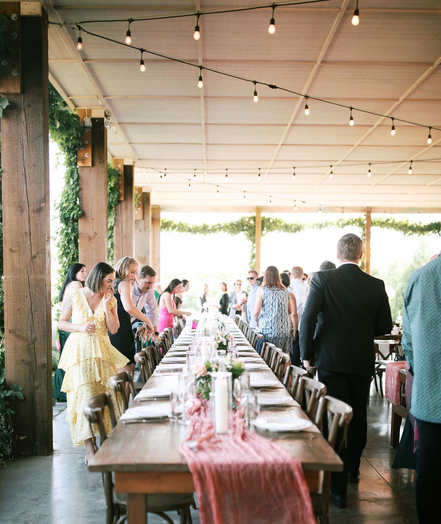 Long rustic dining table with string lights overhead and pink table runner at an indoor-outdoor wedding reception venue.