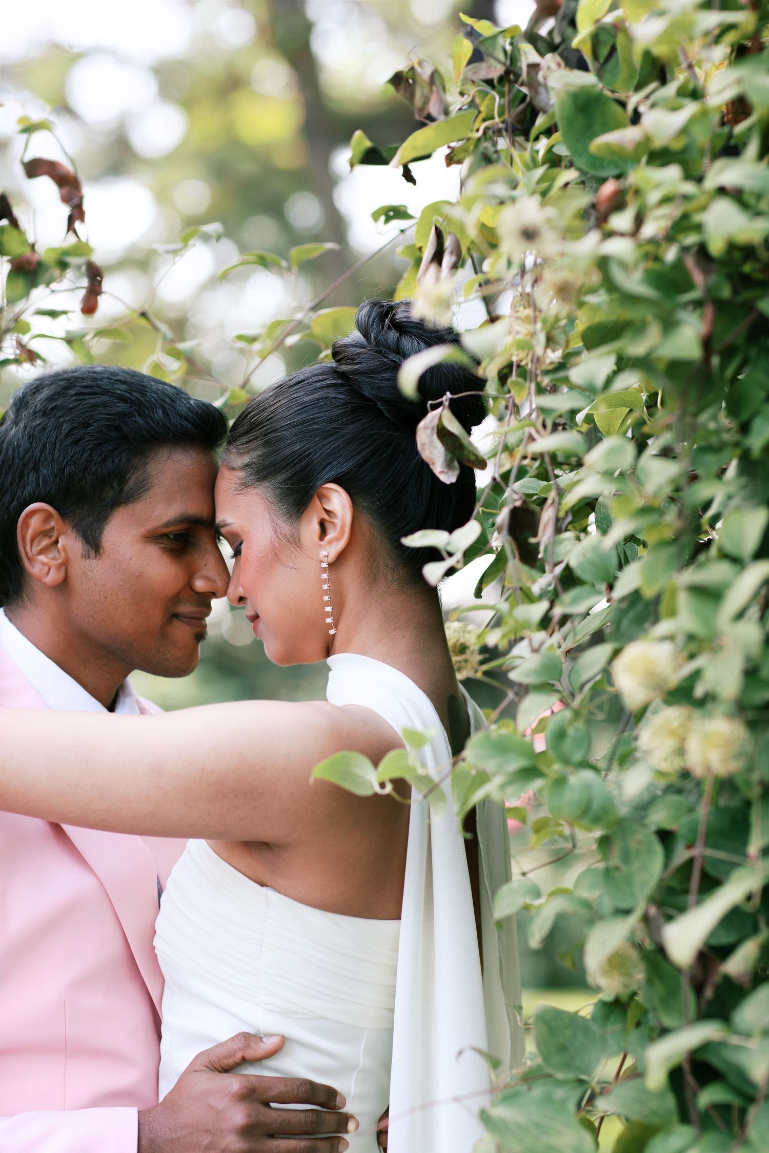 A romantic wedding couple shares an intimate moment in a garden setting with white flowers and greenery.