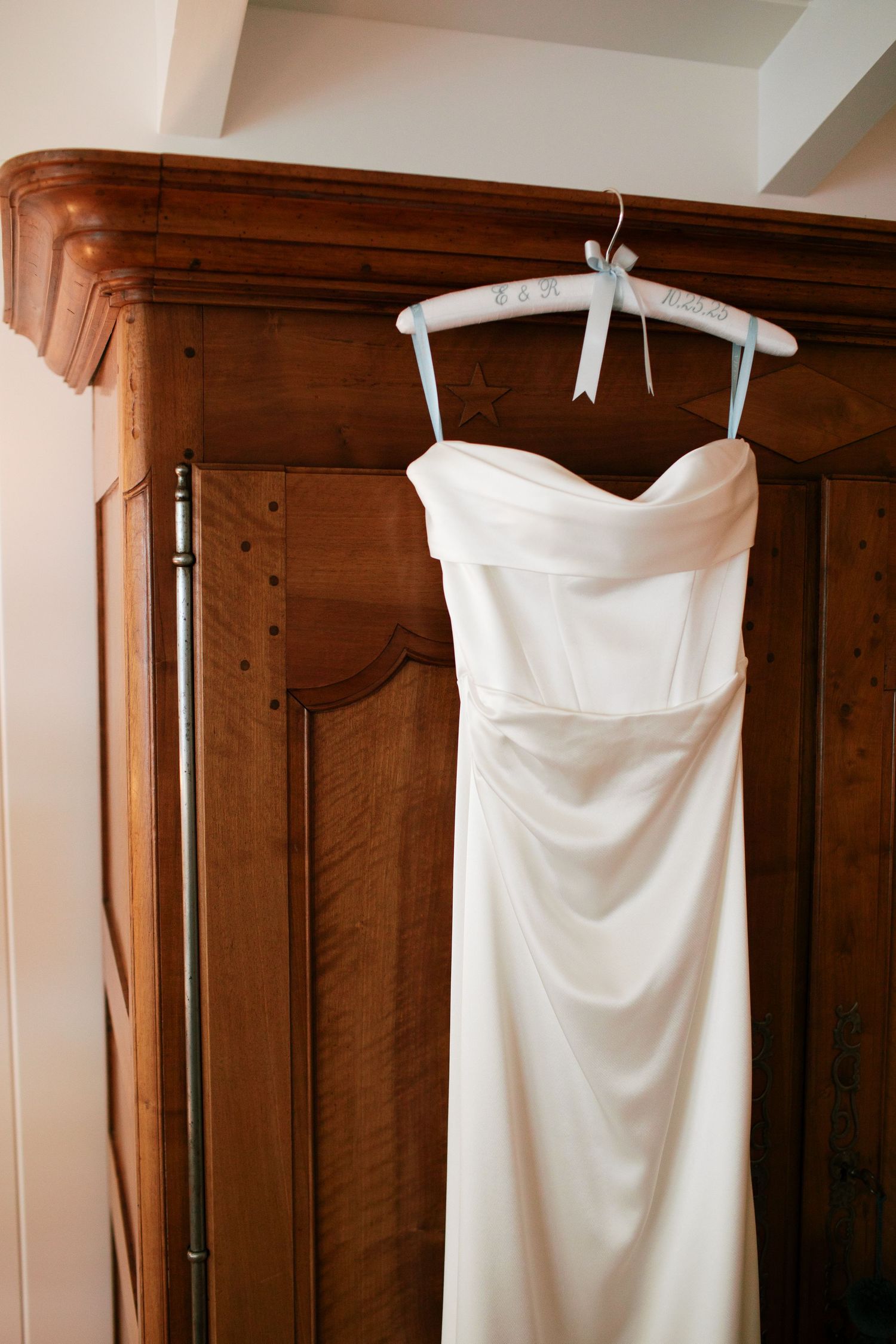 A white strapless wedding dress hangs elegantly against a dark wooden wardrobe in soft lighting.