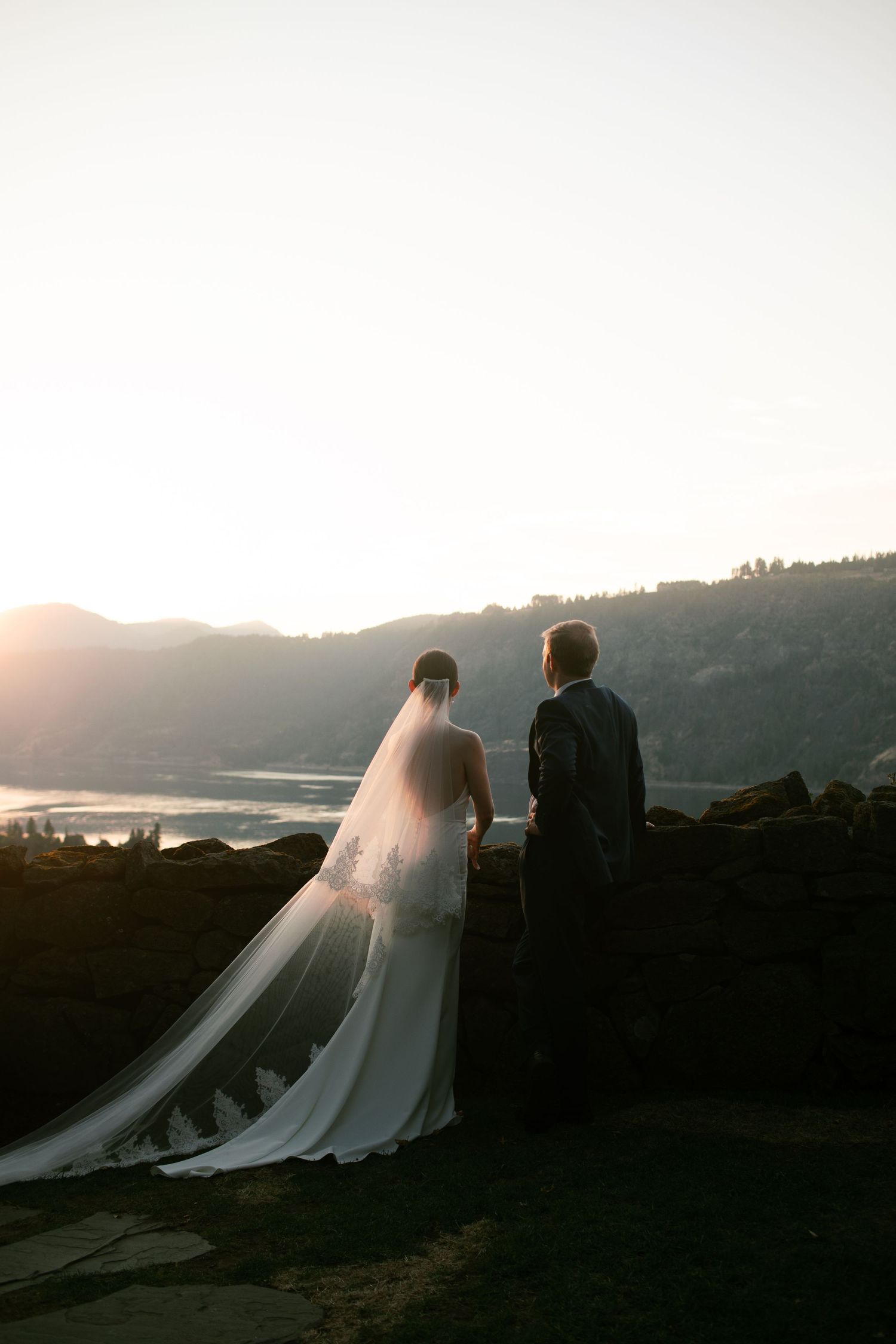 A bride and groom stand silhouetted against a sunset over mountains, with the bride's flowing veil catching the golden light in Hood River.