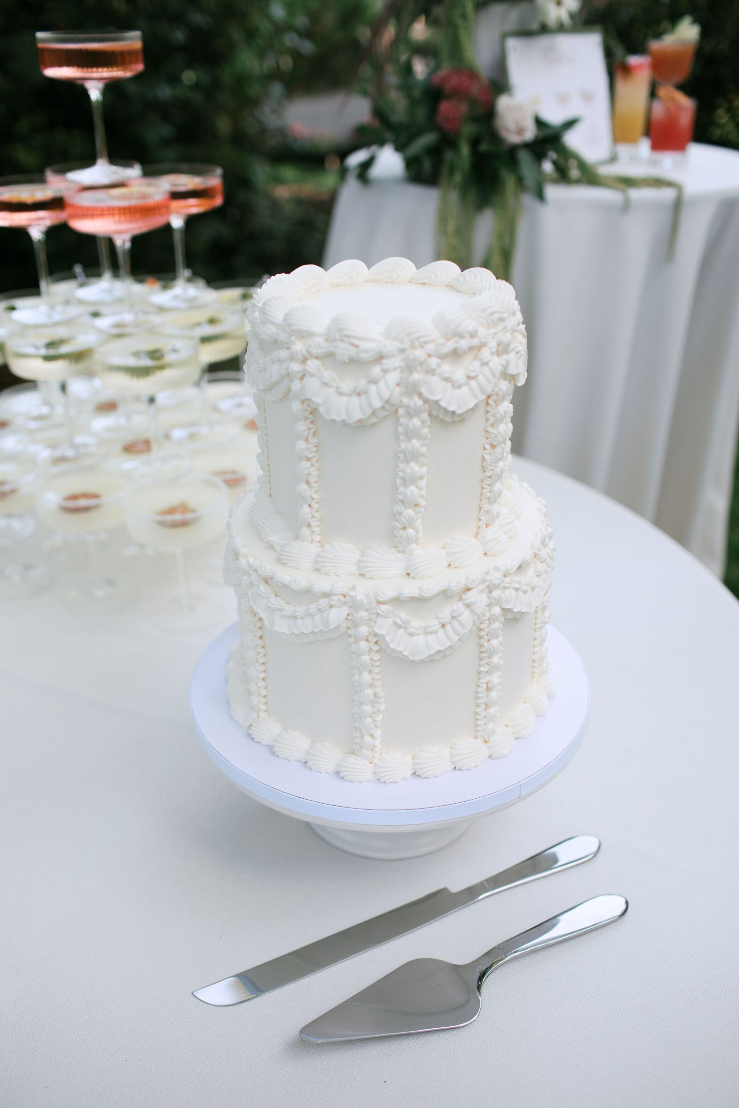A white wedding cake with delicate pearl details sits on a table with serving utensils.