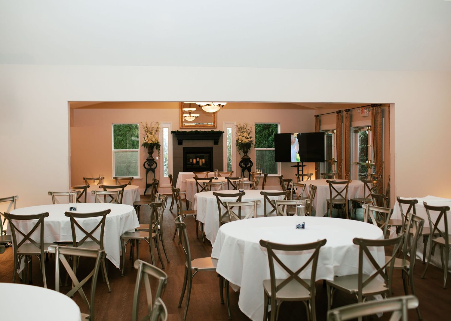 A banquet hall with round tables covered in white tablecloths and wooden cross-back chairs arranged for an event.