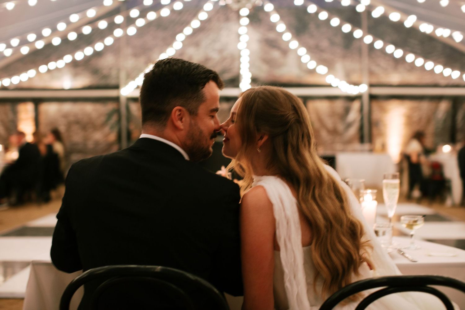 A couple shares an intimate moment under twinkling lights at their wedding reception in a rustic tent venue.