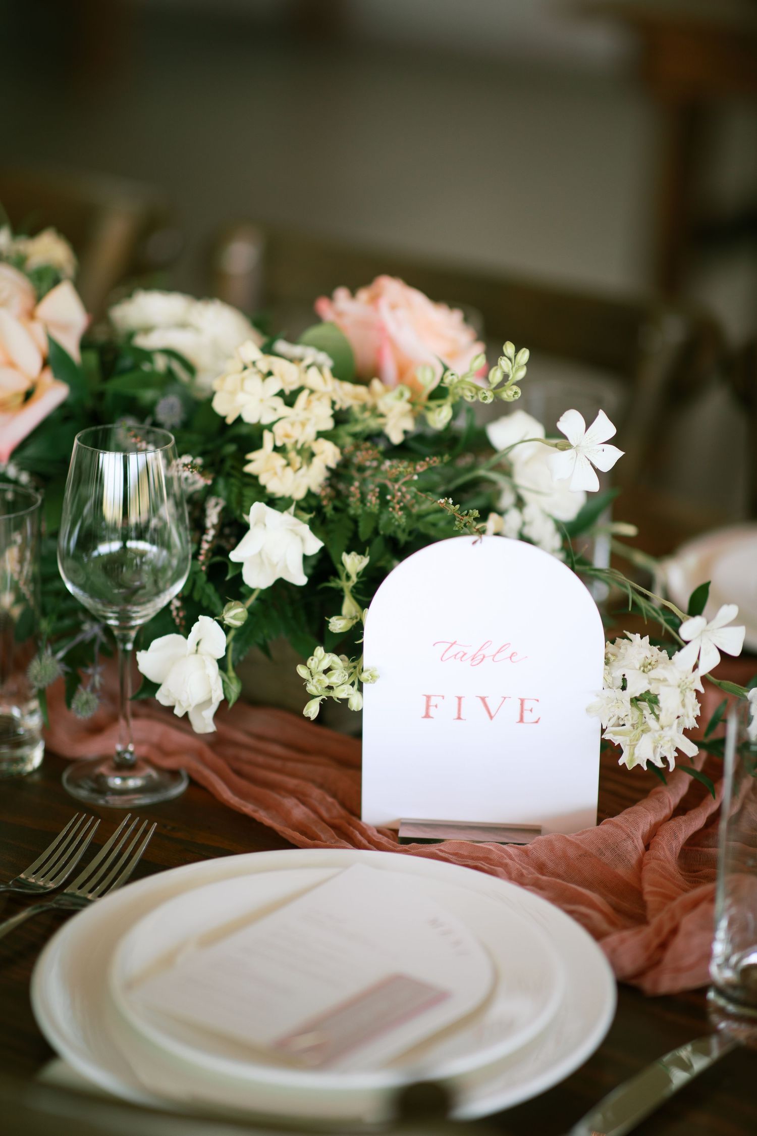 Elegant table setting with pink and white flowers, wine glass, and table number five card on dusty rose fabric.
