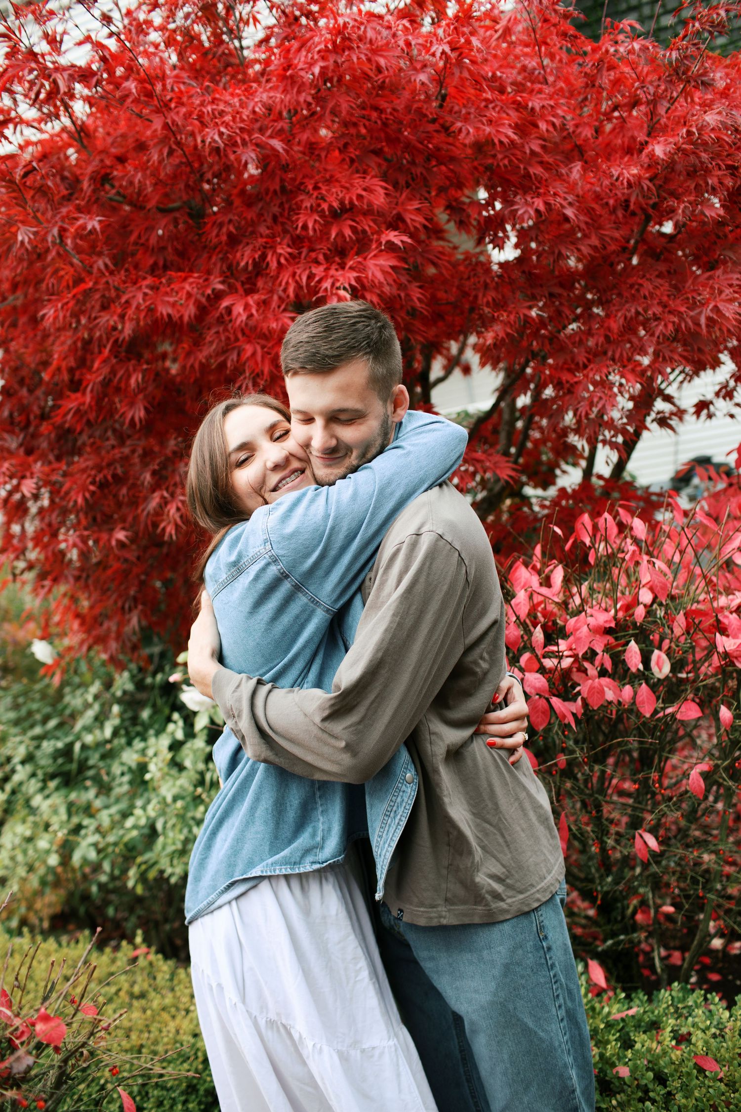 A couple embraces under vibrant red autumn maple leaves in a romantic outdoor setting.