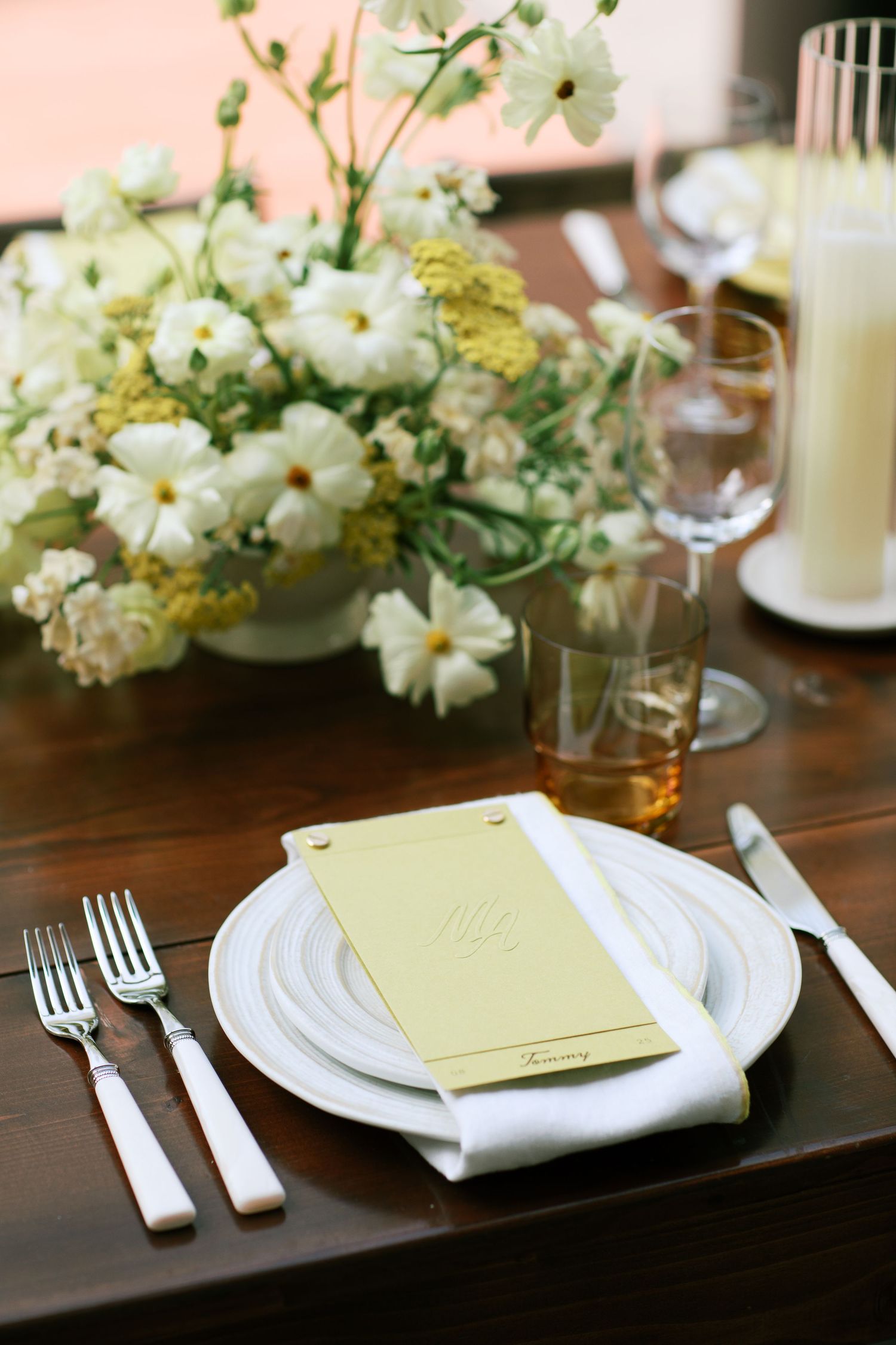 Elegant place setting with white flowers, napkin and yellow menu card on dark wooden table at formal dinner event.