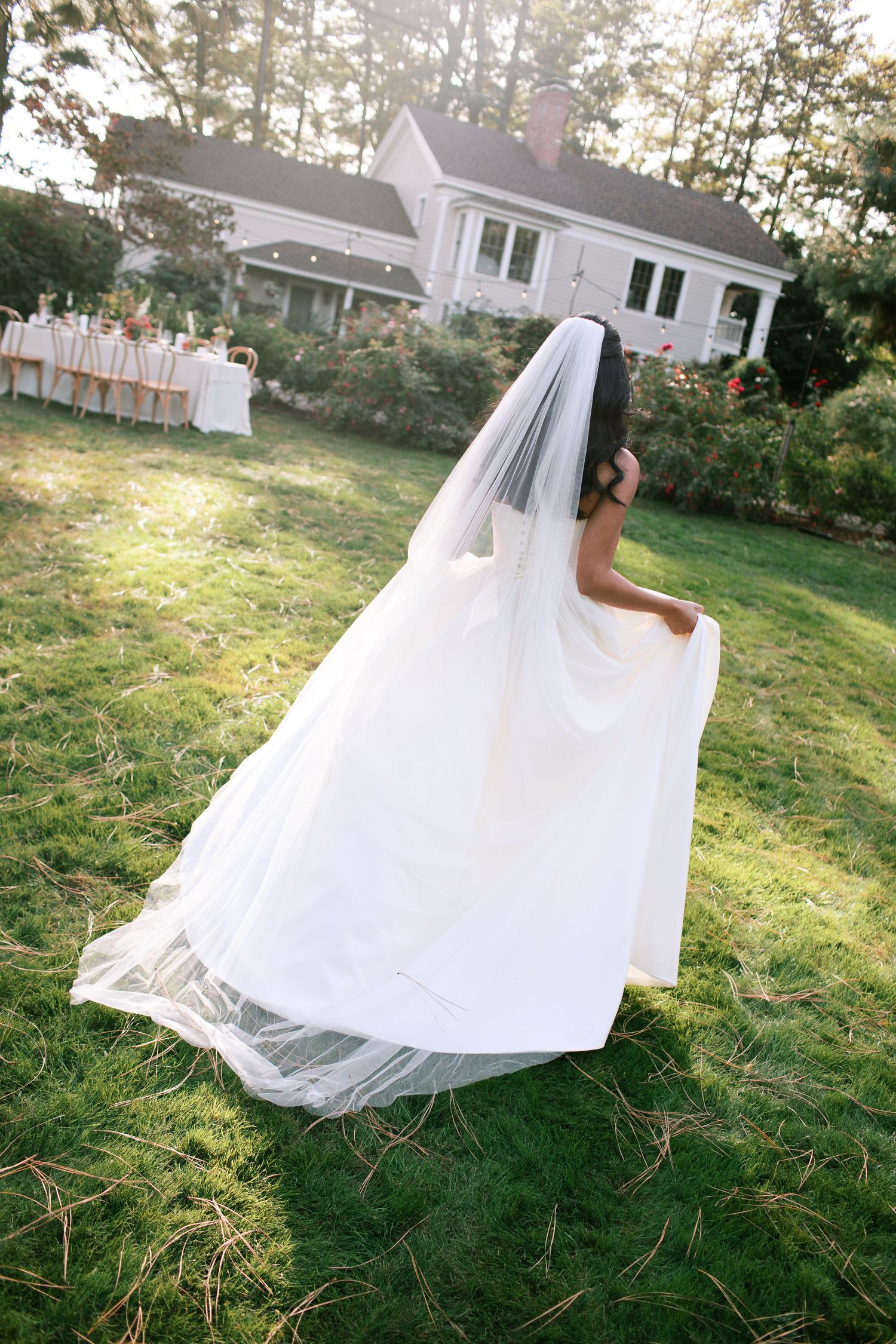 A flowing white wedding dress billows in the wind as it moves across a sunlit lawn on a summer afternoon.