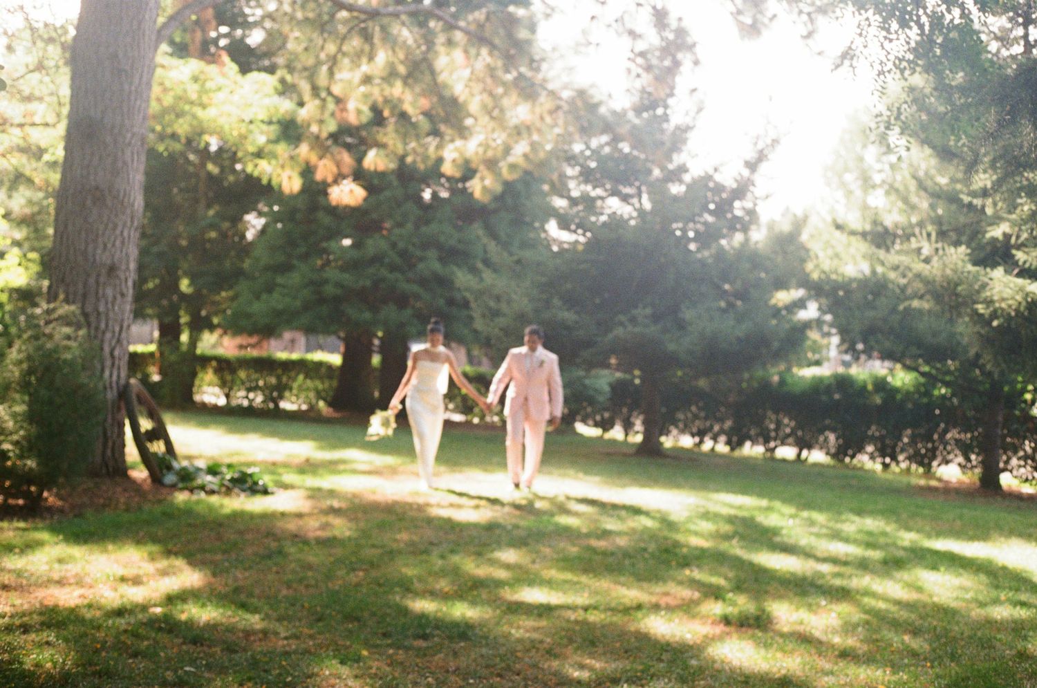 A couple walks hand in hand through a sunlit park with trees casting long shadows on the grass.