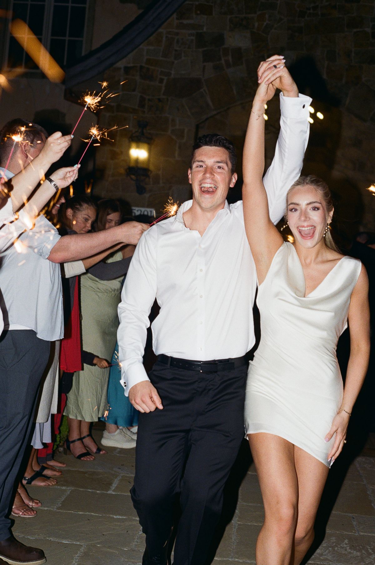 Joyful wedding guests dancing energetically in formal attire at an evening reception with stone walls and warm lighting.