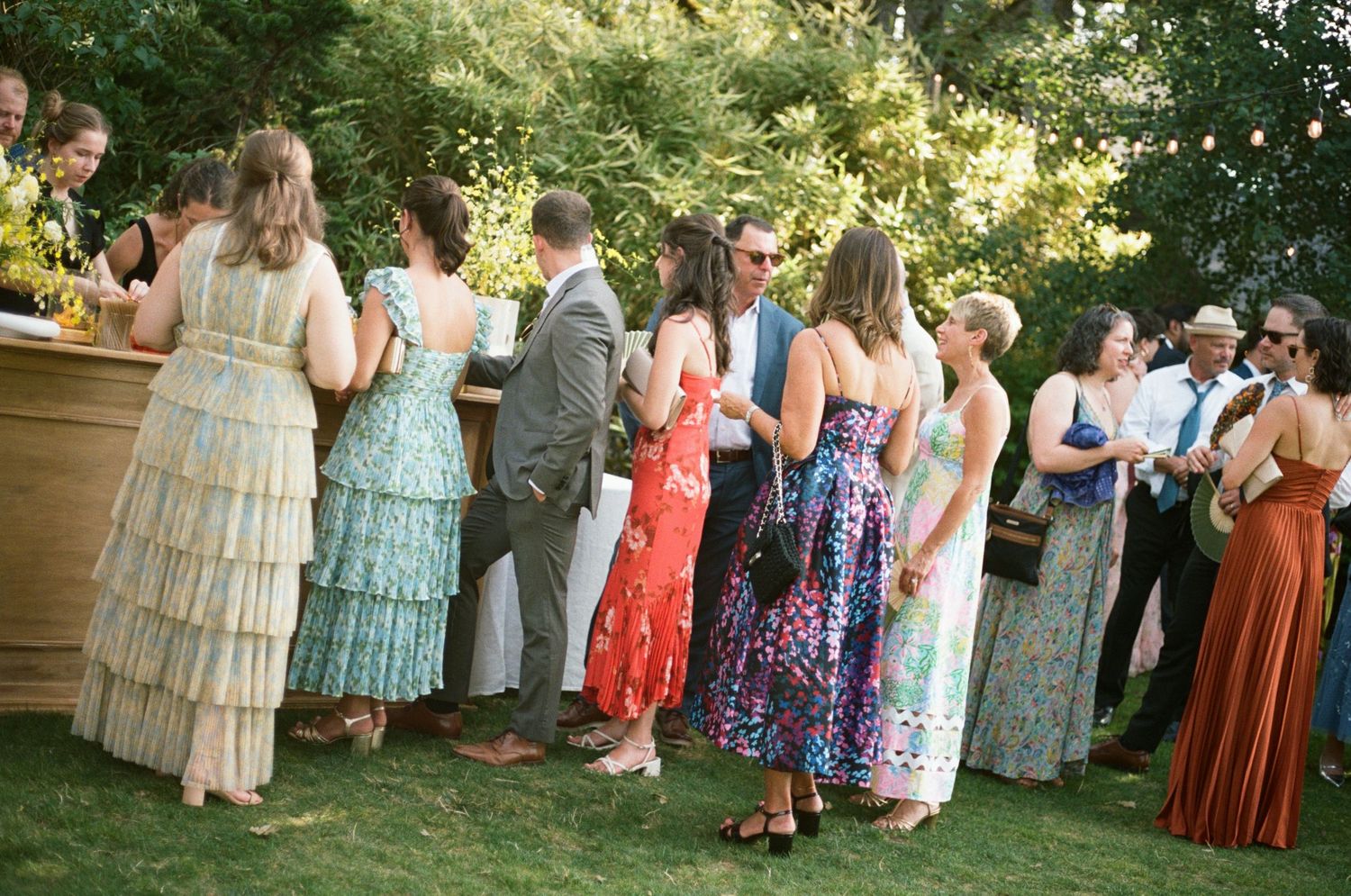 Wedding guests in colorful dresses and formal attire gather outdoors in a garden setting during a summer celebration.