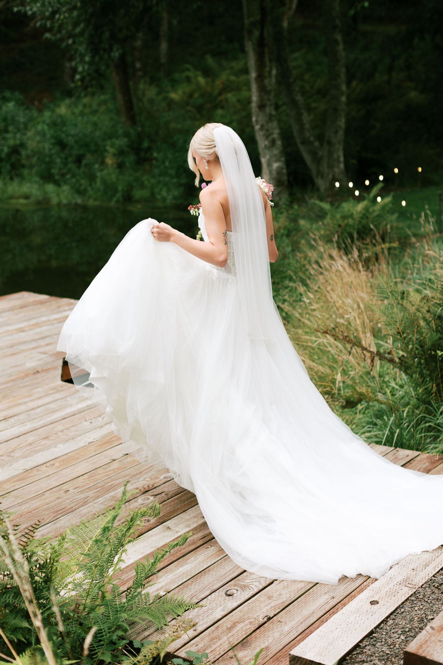 A flowing white wedding dress with a long veil twirls gracefully on a wooden deck surrounded by lush greenery.