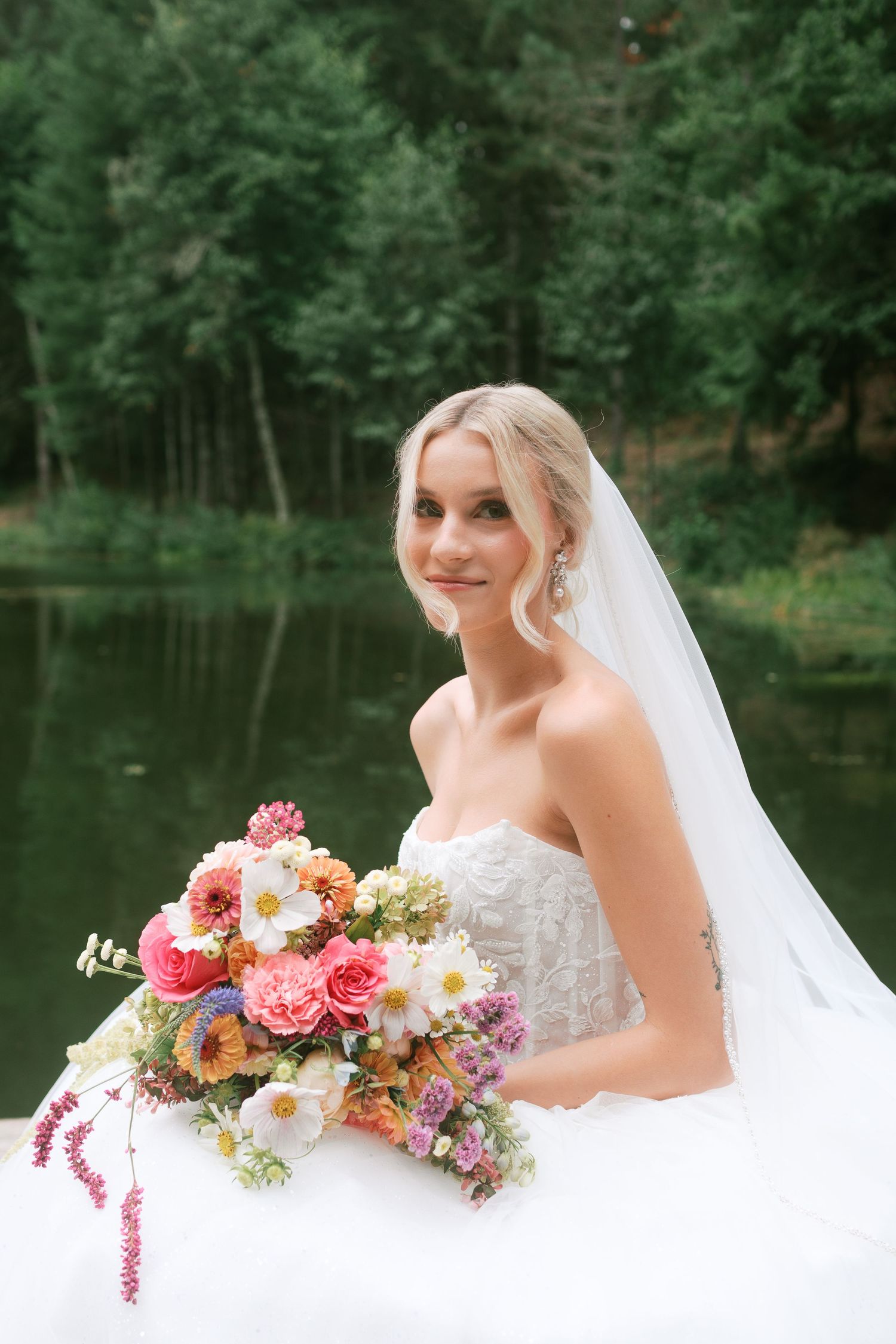 A romantic lakeside wedding scene with a beautiful floral bouquet featuring pink and white blooms against a white dress.