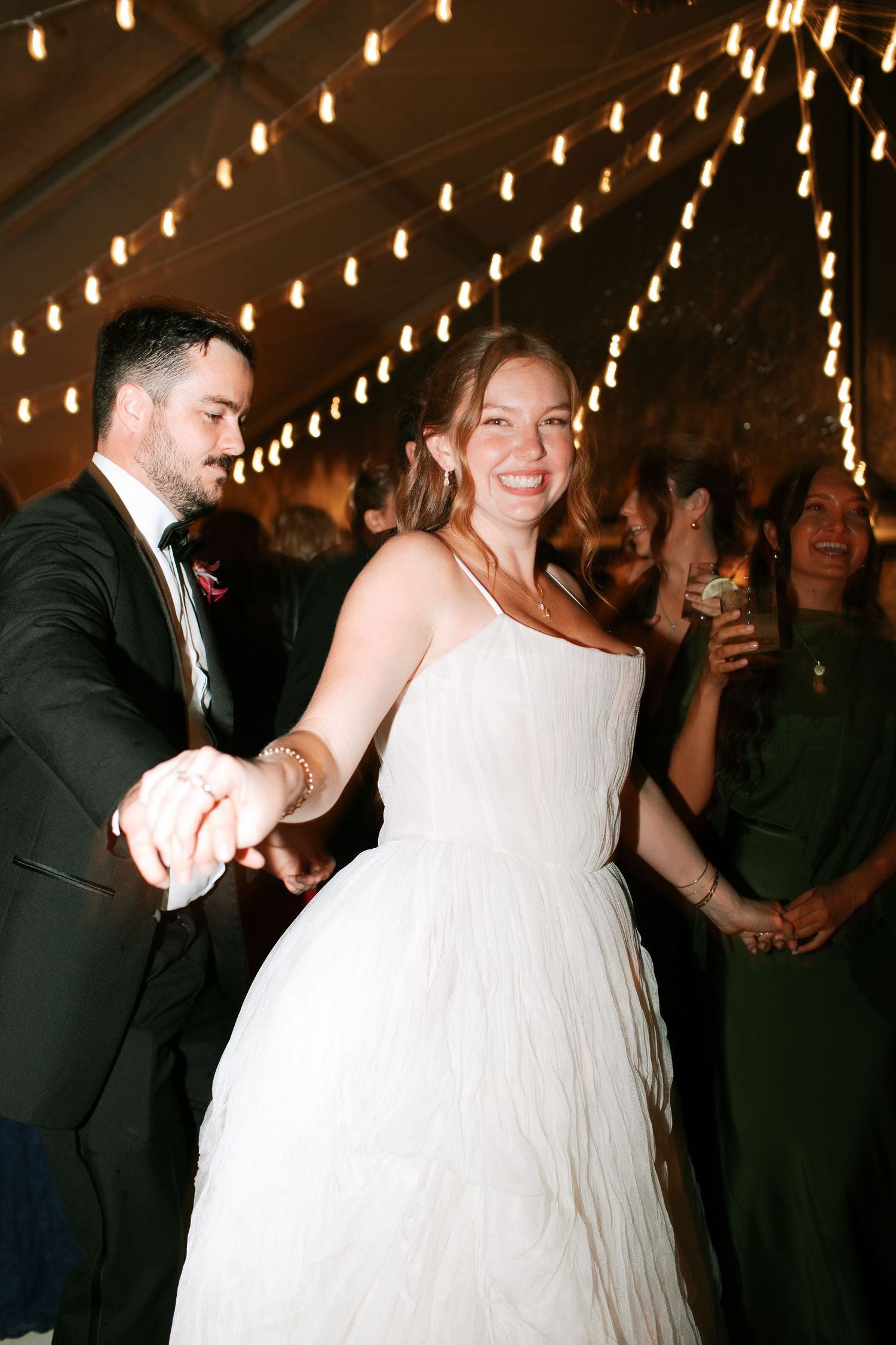 A couple dances together at a wedding reception under romantic string lights while guests watch in the background.