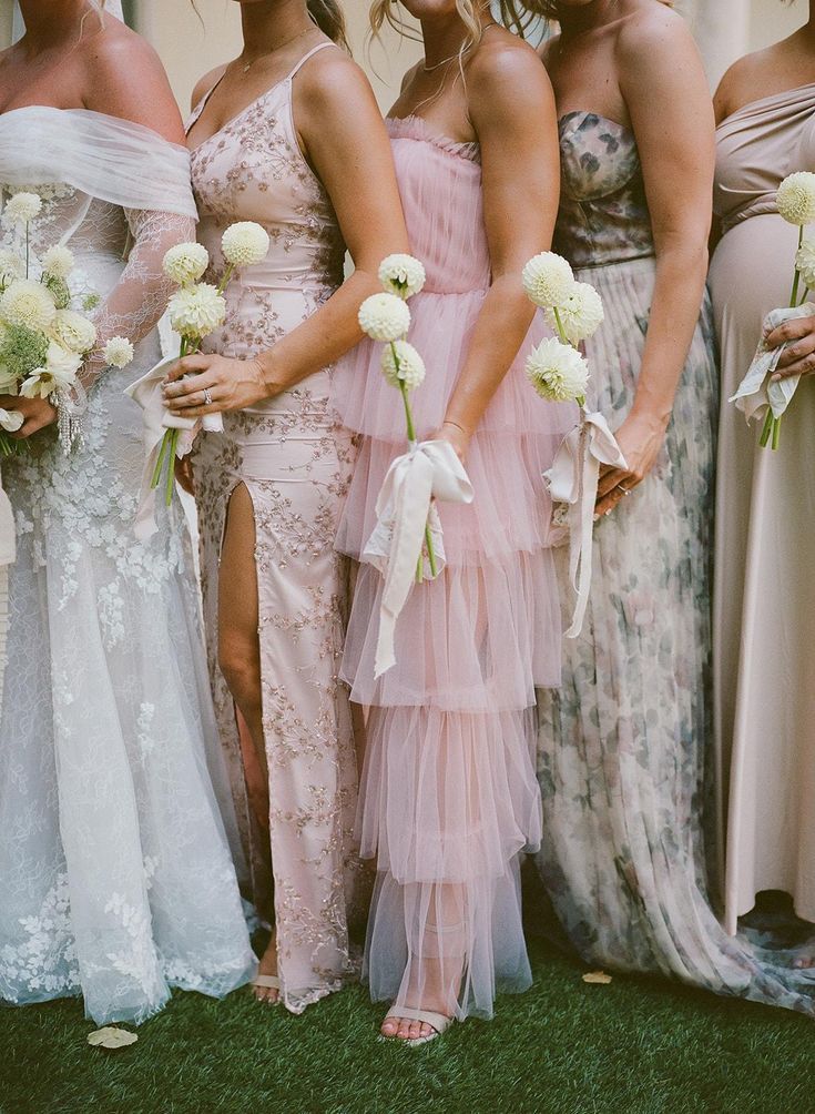 Bridesmaids in soft pink and white lace dresses hold delicate white floral bouquets while standing on grass.