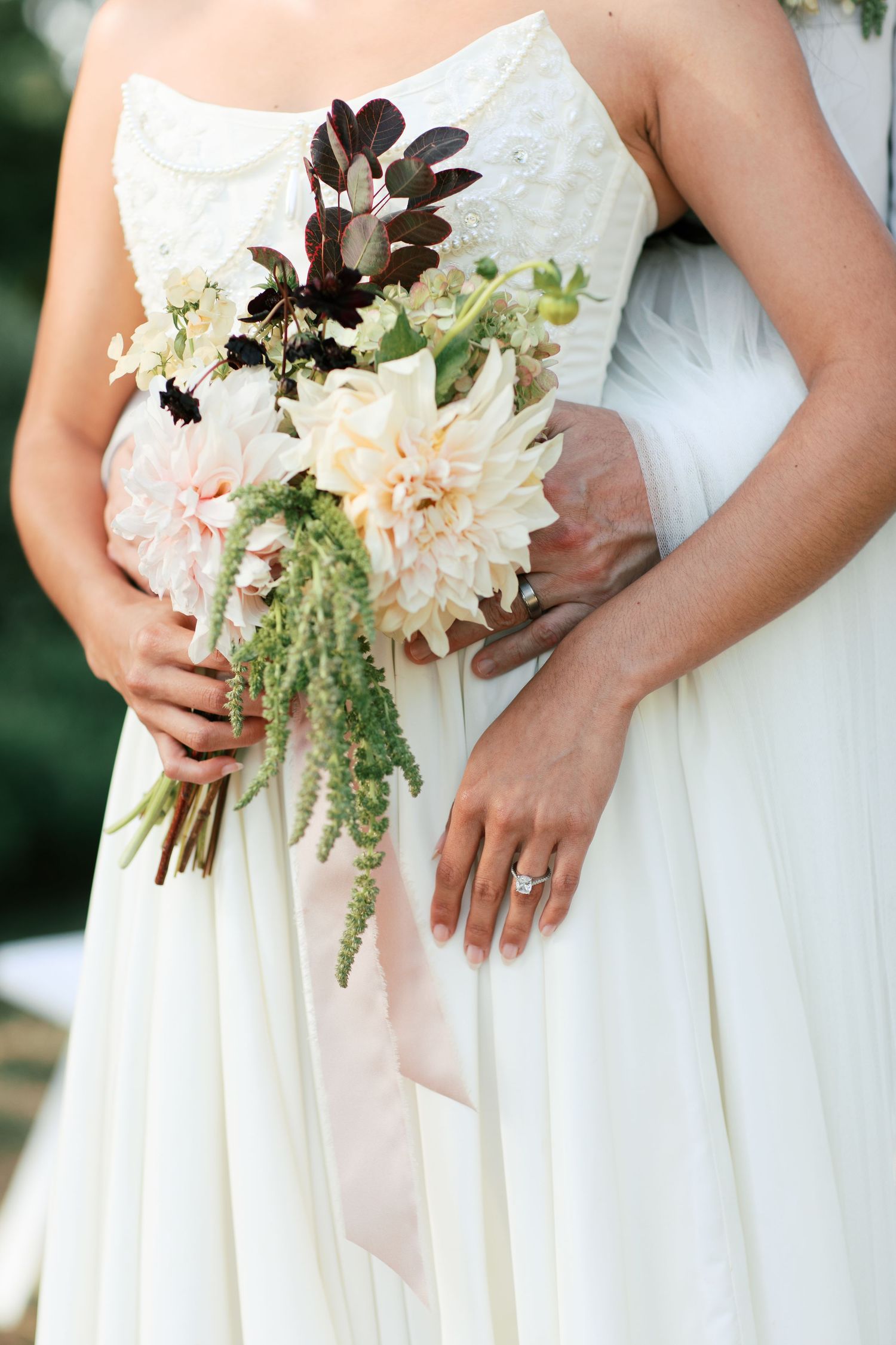 Row of elegant white bridal bouquets featuring soft pink dahlias, greenery and dark foliage against ivory dresses.