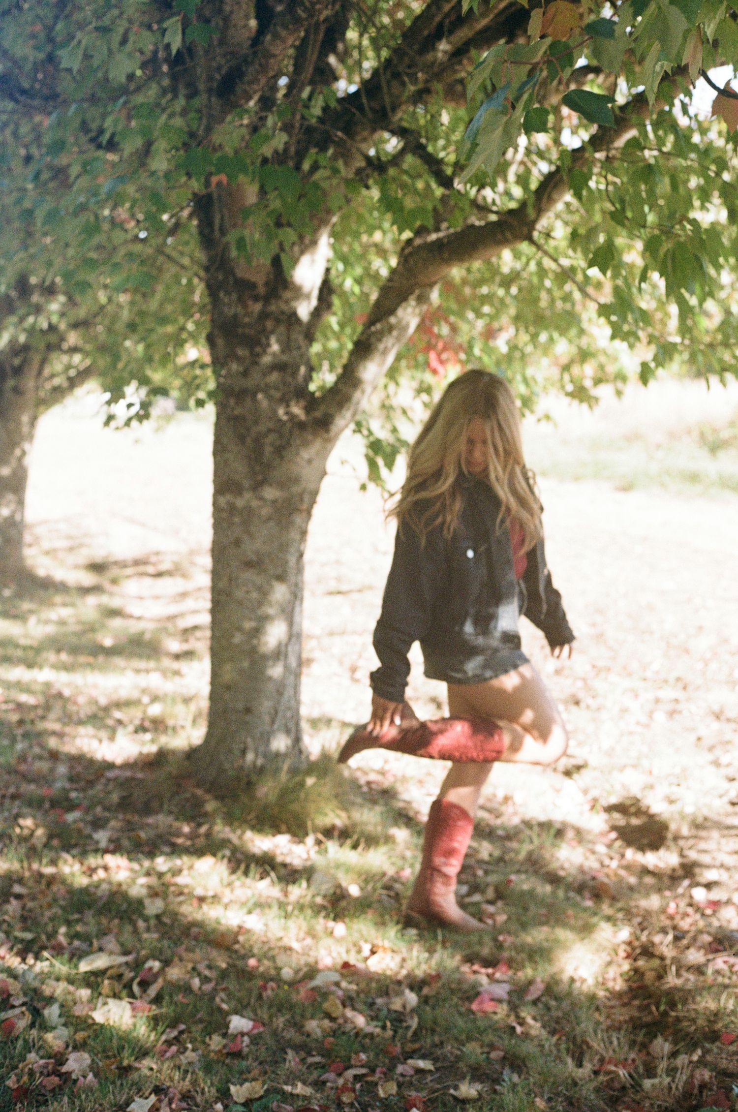 Dappled sunlight illuminates red boots walking through fallen leaves under orchard trees on 35mm film-Adrianna Elizabeth Photography.