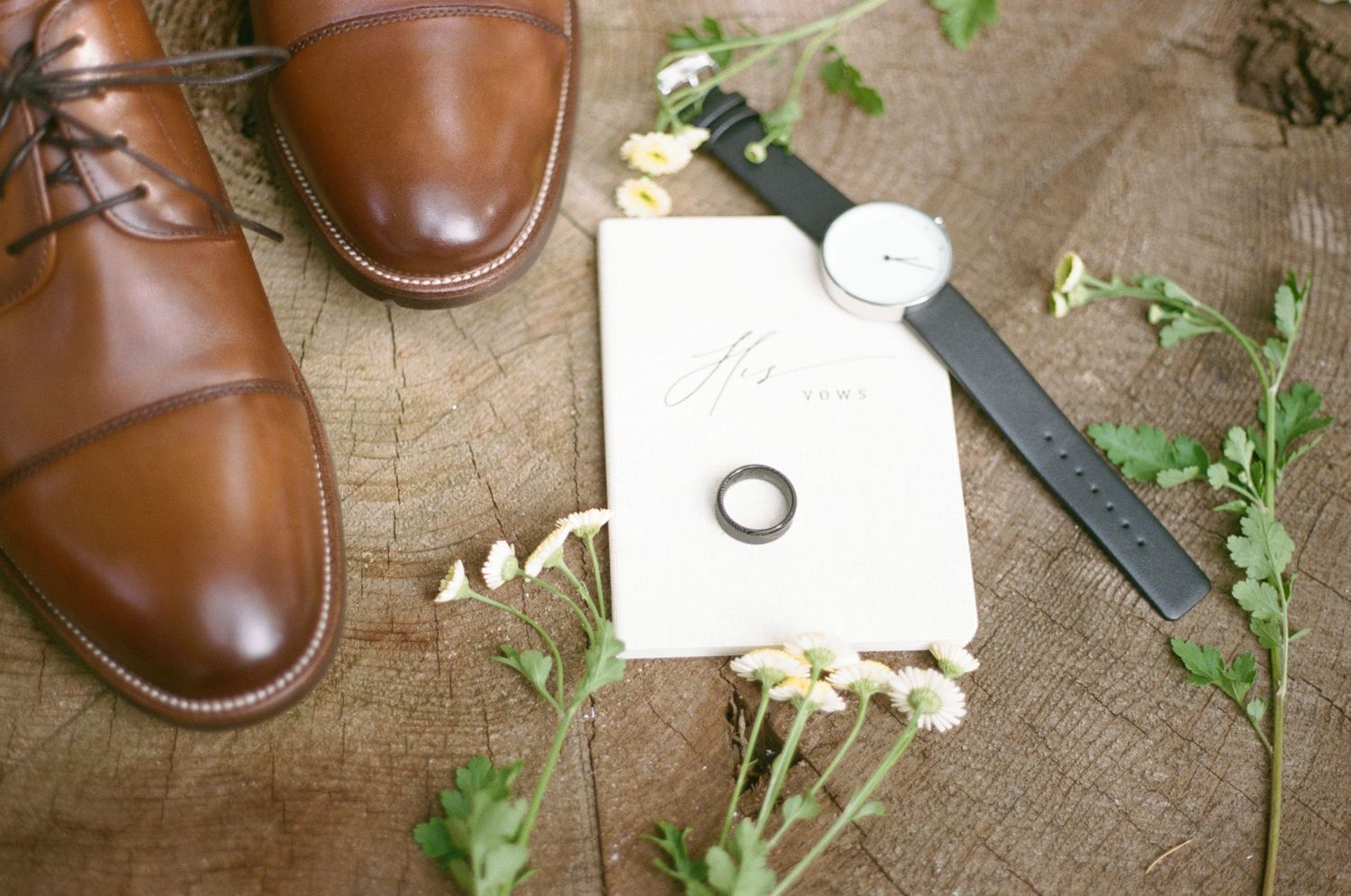 Polished brown leather shoes arranged with green plant sprigs and styling accessories on rustic wooden surface on 35mm film.