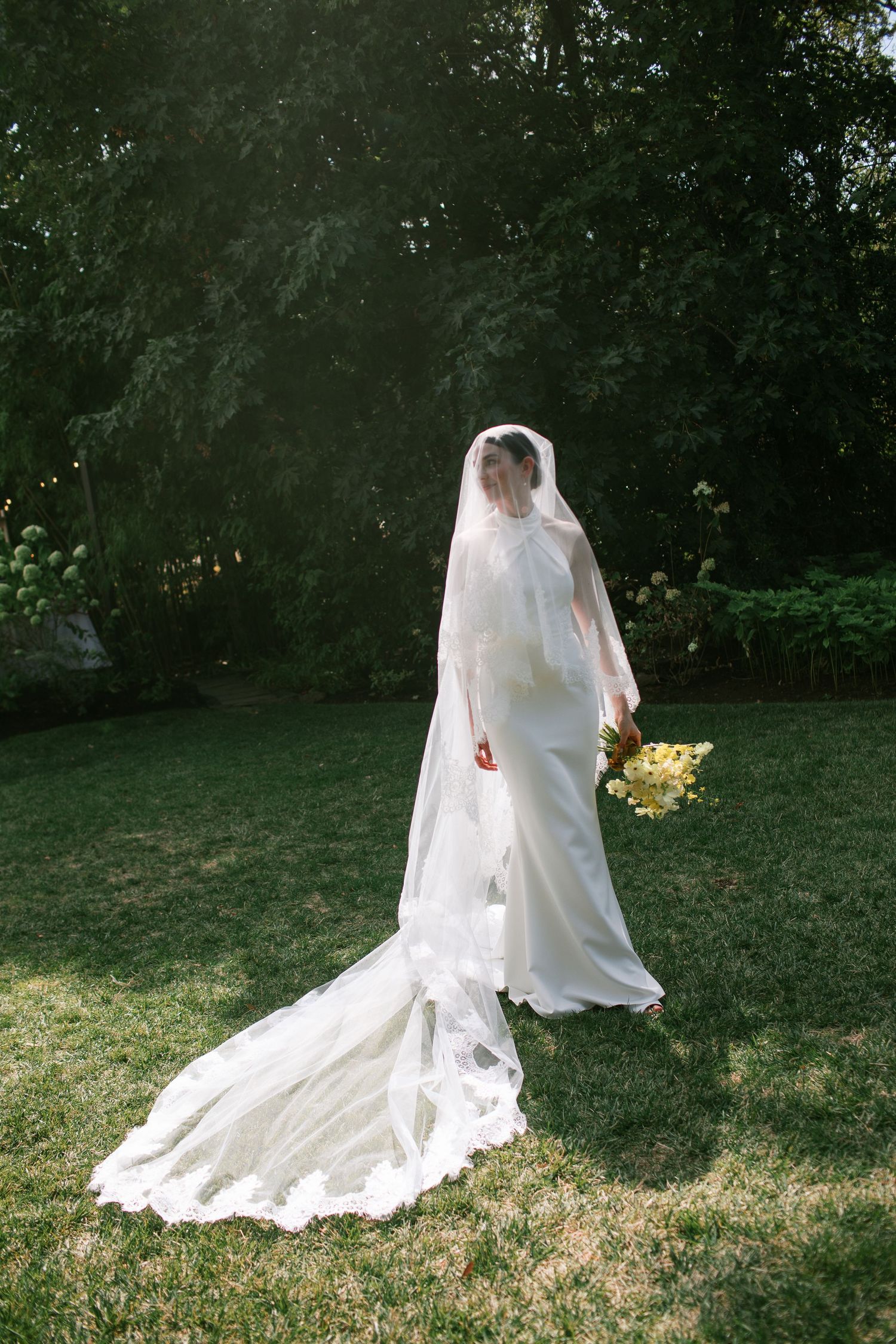 A bride in a flowing white wedding gown and veil sits gracefully on grass in a garden setting.
