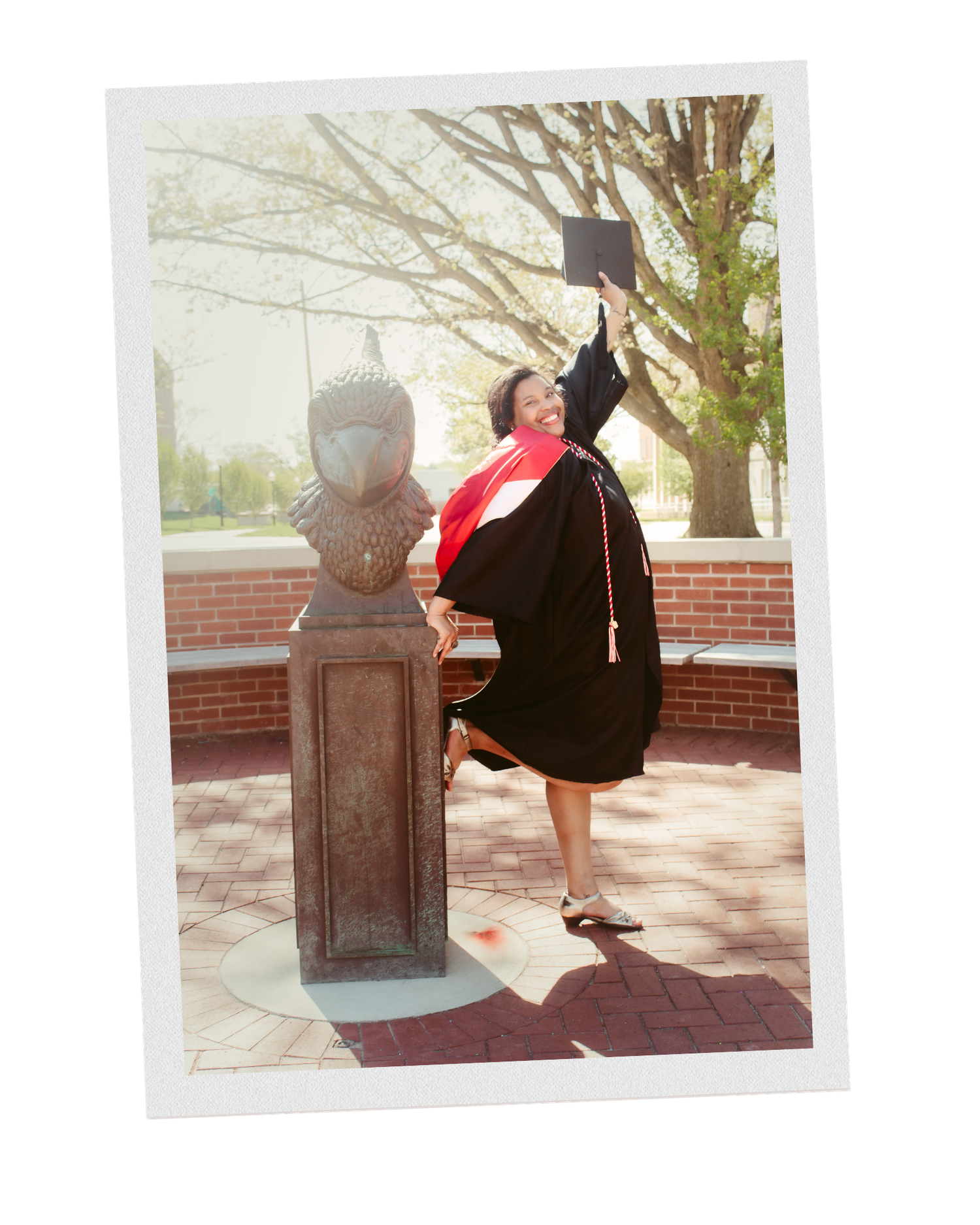 Senior graduating from college, holding their cap in the air and leaning on the ISU Redbird statue while wearing a gown