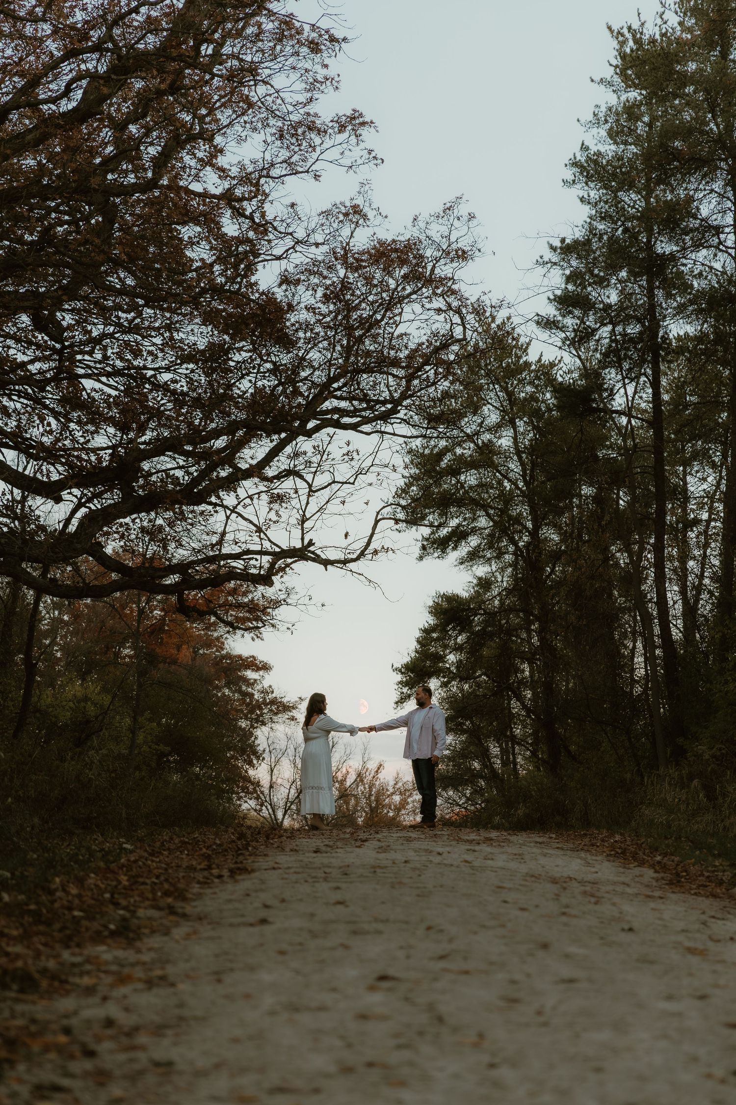 Couple cinematically reaches toward one another while the moon sits on top of their arms during their engagement session