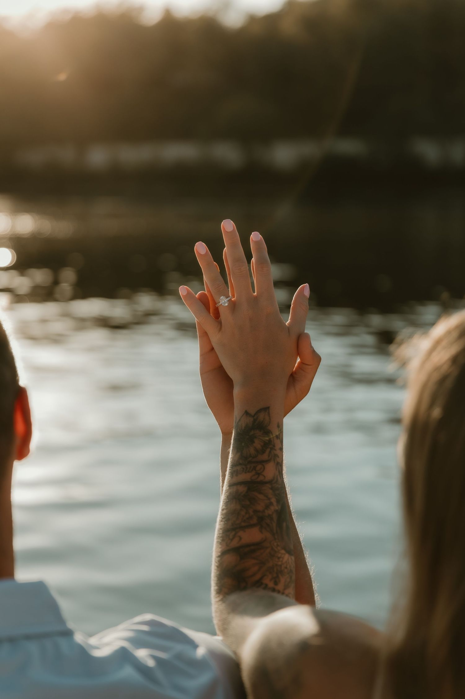 Close-up of couple’s hands showing off the engagement ring. Romantic outdoor moment with soft reflections and treeline at sunset.