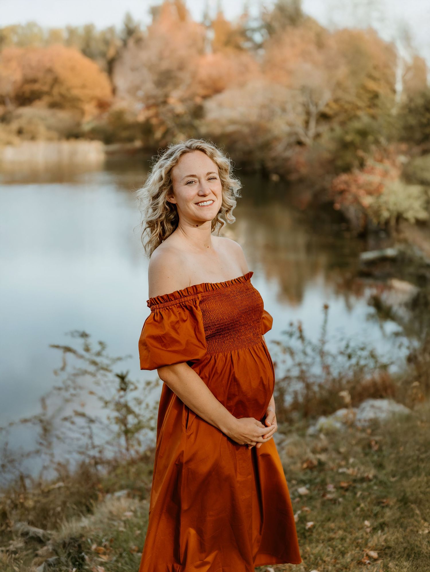 Expecting mom holds her belly in front of fall colors and a lake during a maternity photo session with central il maternity photographer