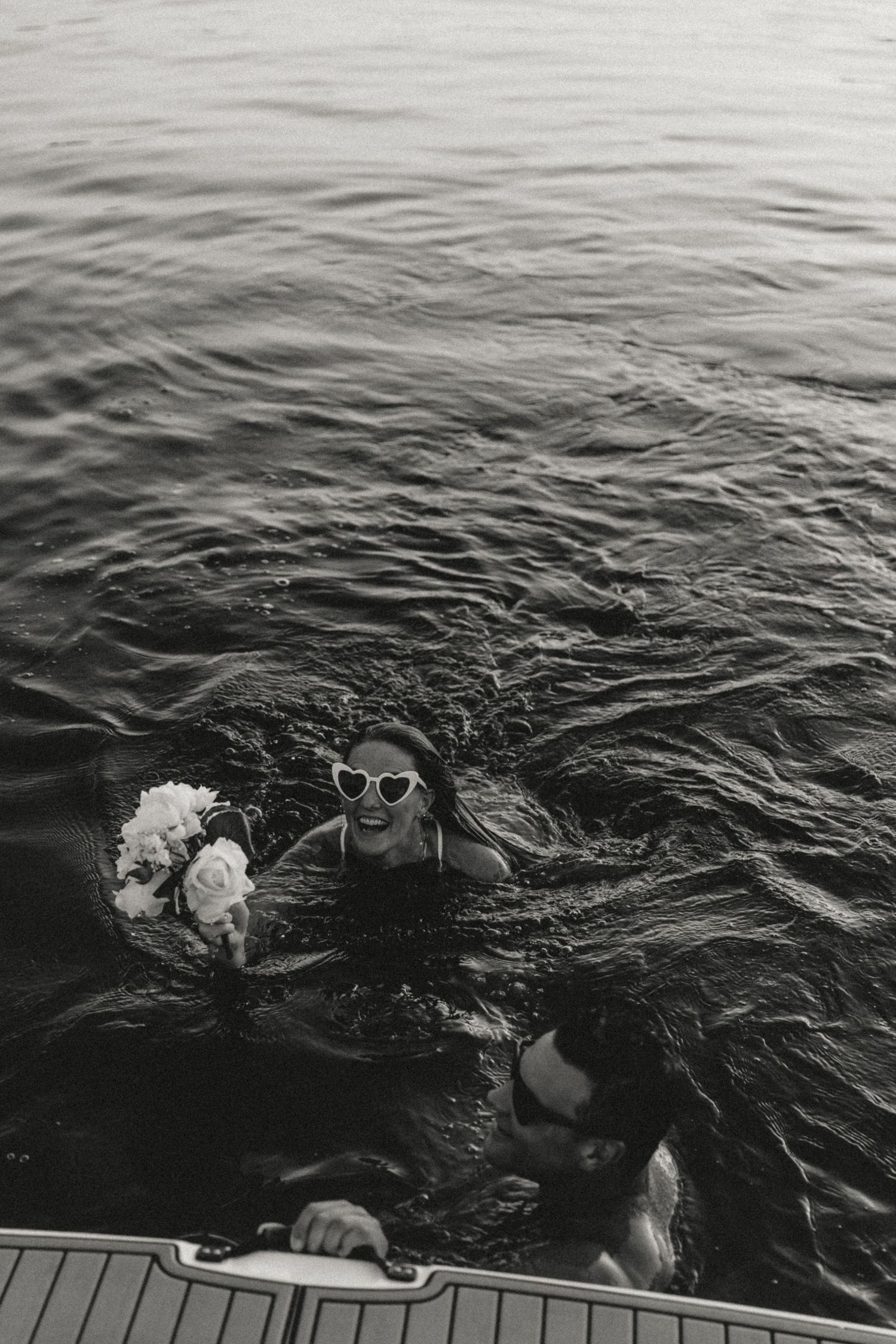 Black-and-white photo of joyful couple swimming near a boat. Playful, romantic moment captured in water, evoking summer love.
